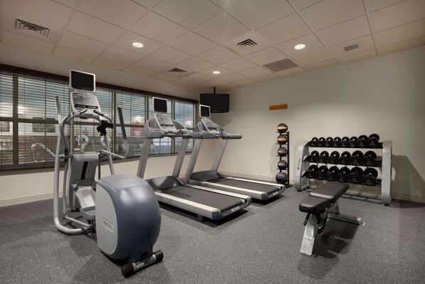 Un gimnasio con cintas de correr, una elíptica, una bicicleta estática y una banca. Se ven pesas sobre un soporte y un televisor en la pared.