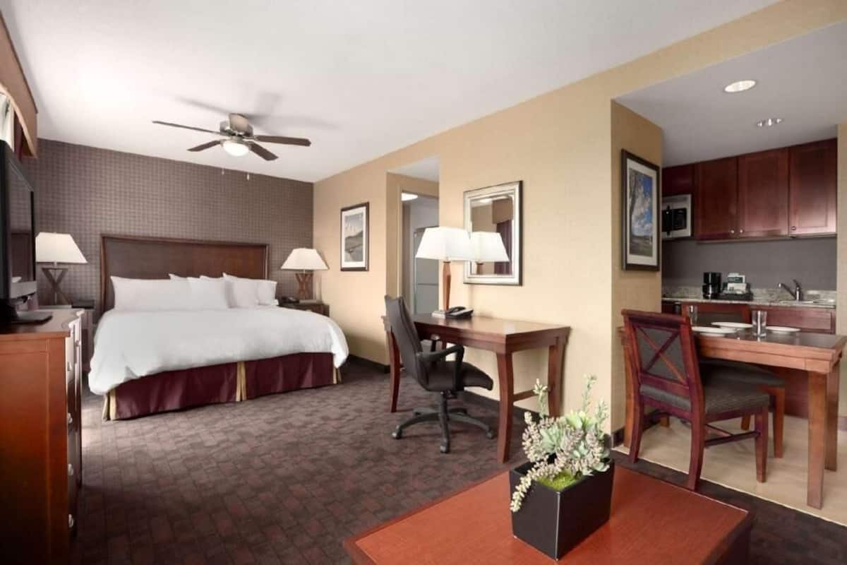 A hotel room with a bed, desk, chair, kitchenette, and a ceiling fan.