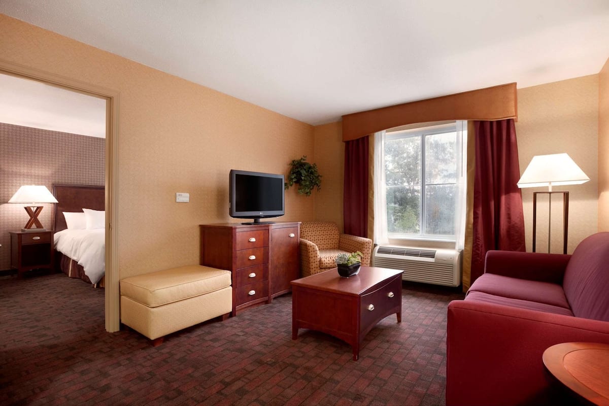 A hotel room with a bed, a TV, a desk, a chair, a sofa, and a window with curtains.