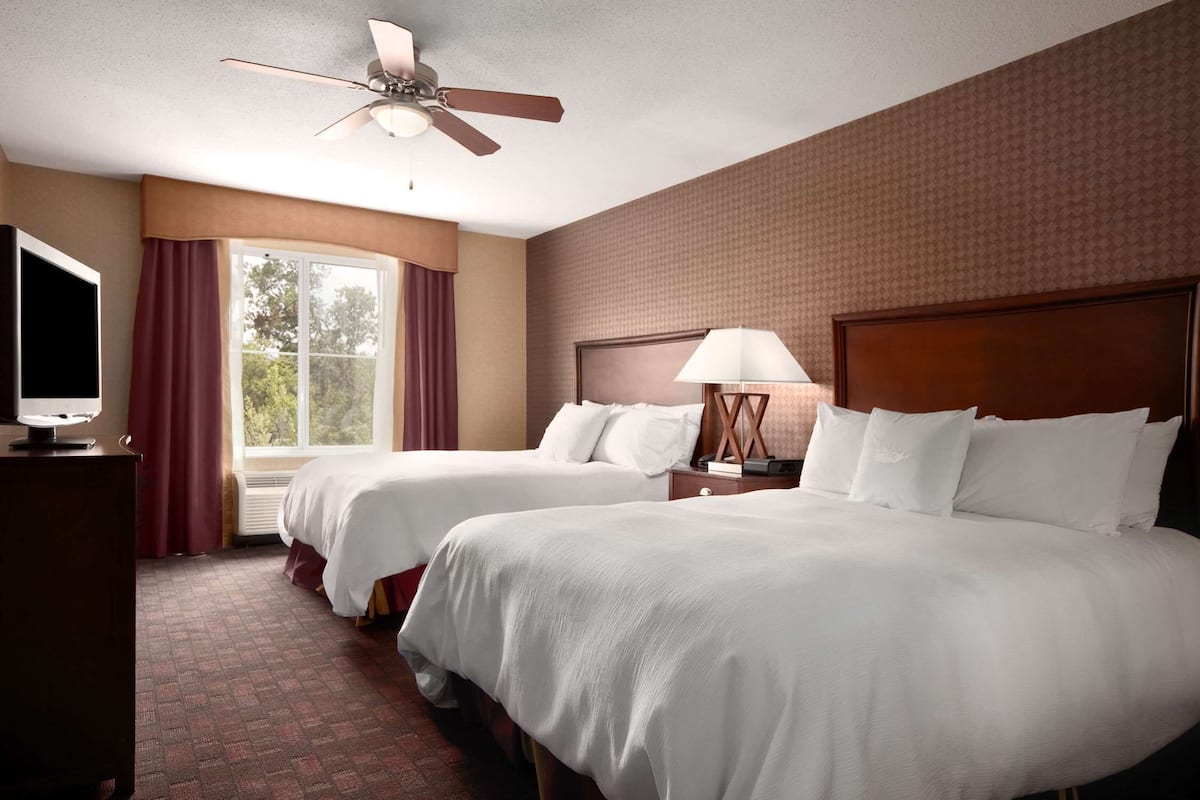 A hotel room with two beds, a ceiling fan, a TV, and a window with curtains.