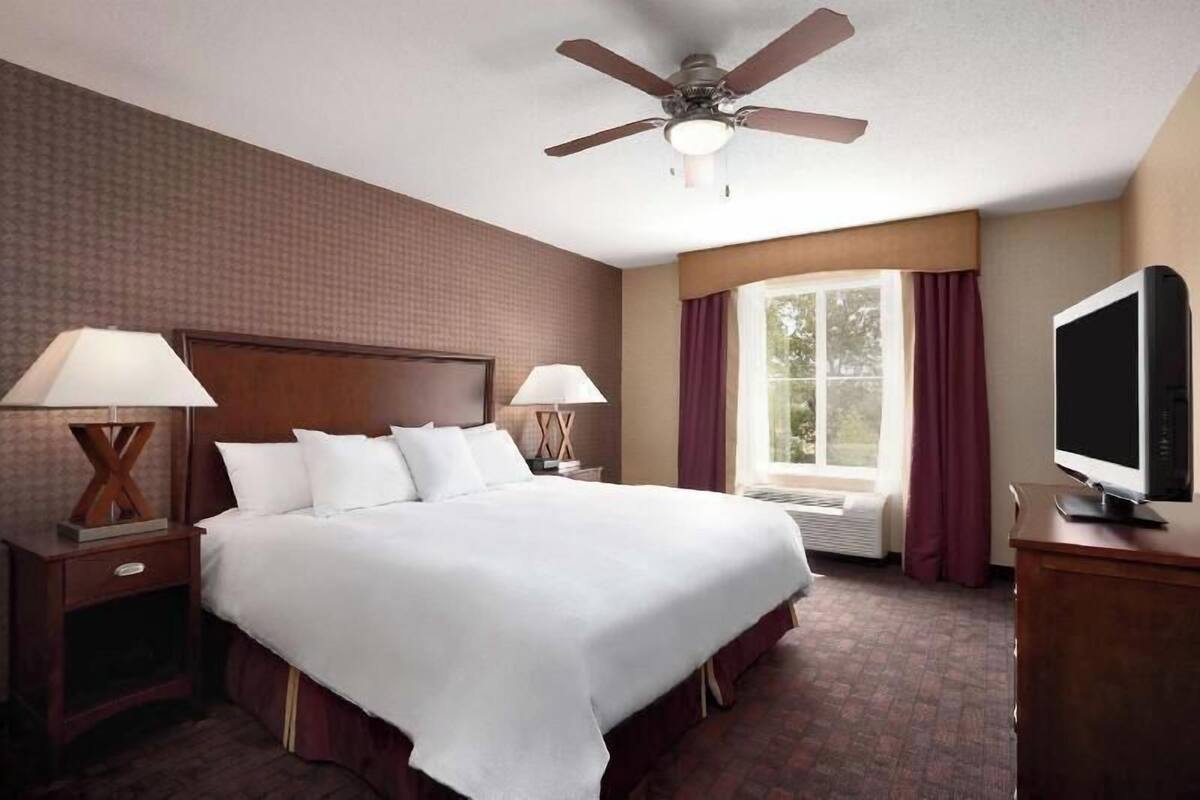 A hotel room with a bed, two bedside lamps, a ceiling fan, a TV, a window with curtains, and a radiator.