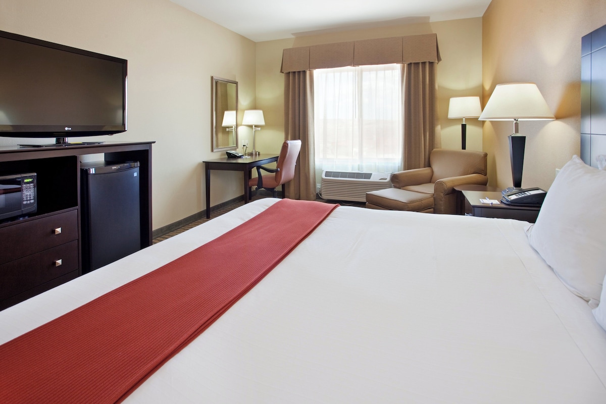 A hotel room with a bed, a desk with a TV, a chair, a lamp, and an air conditioning unit.