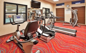 Un gimnasio con cintas de correr, elípticas y una bicicleta estática, con piso cubierto de alfombra roja y un reloj en la pared.
