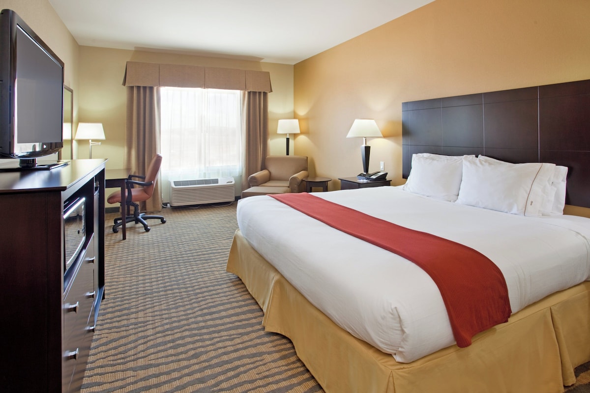 A hotel room with a large bed, a desk with a television, two chairs, a desk lamp, and a window with curtains.
