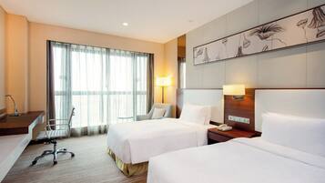 A hotel room with two beds, a desk, a chair, a lamp, and a large window with curtains.