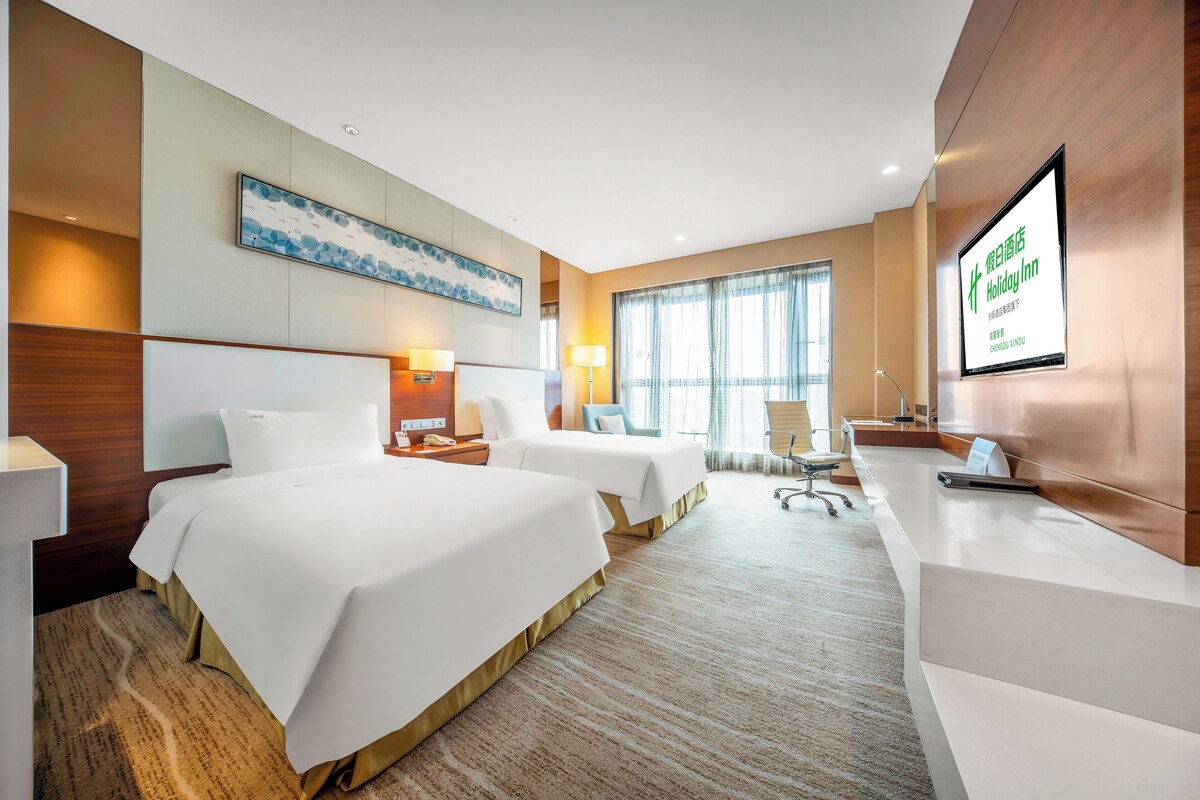 A hotel room with two beds, a desk, a chair, a TV, and a large window.