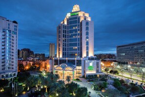A multi-story hotel with a prominent Holiday Inn sign, surrounded by other buildings and a well-maintained landscaped area with trees and shrubs.