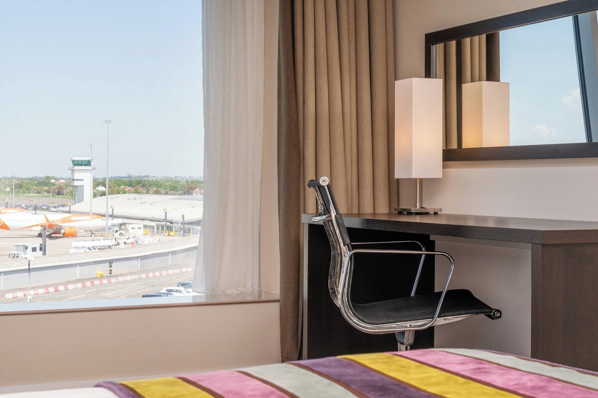 A hotel room with a bed, a desk, a chair, a window with a view of an airport, and a lamp.