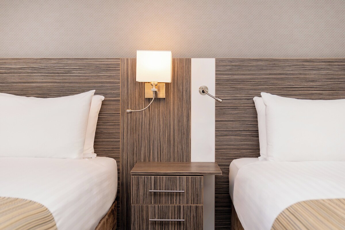 A hotel room with two beds, a nightstand, a wall-mounted lamp, and a towel rack.
