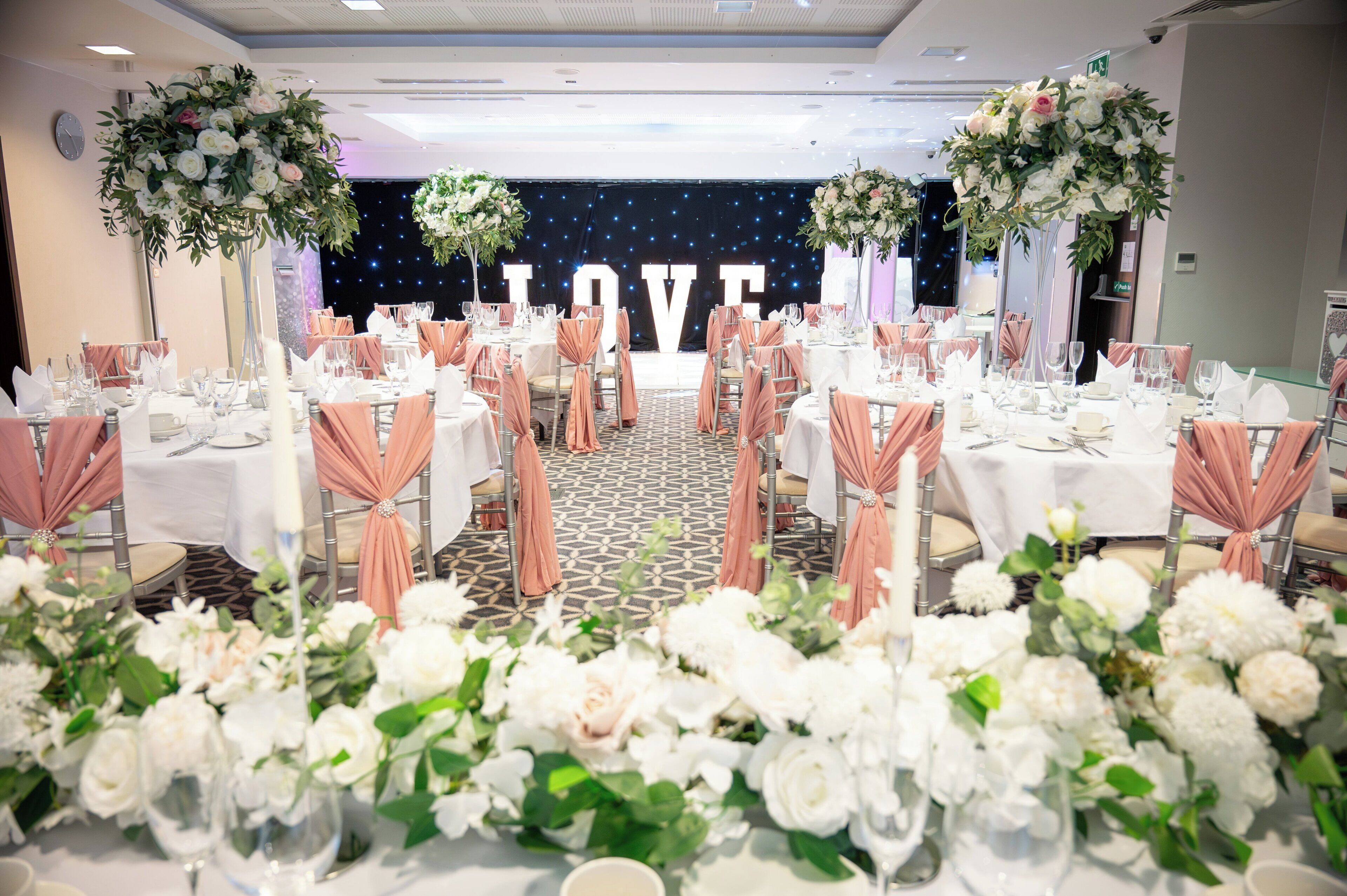 Une salle de banquet avec des tables rondes, des housses de chaise de couleur pêche et des compositions florales blanches.