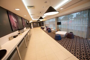 Un hall d’hôtel moderne avec un long comptoir d’accueil, un espace salon agrémenté de tabourets colorés, de grands luminaires suspendus et un tapis à motifs.