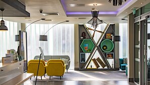 A modern reception area with a desk, two yellow chairs, a grey sofa, and a unique geometric shelving unit with books and decorative items. - Holiday Inn Southend by IHG (Southend-on-Sea)