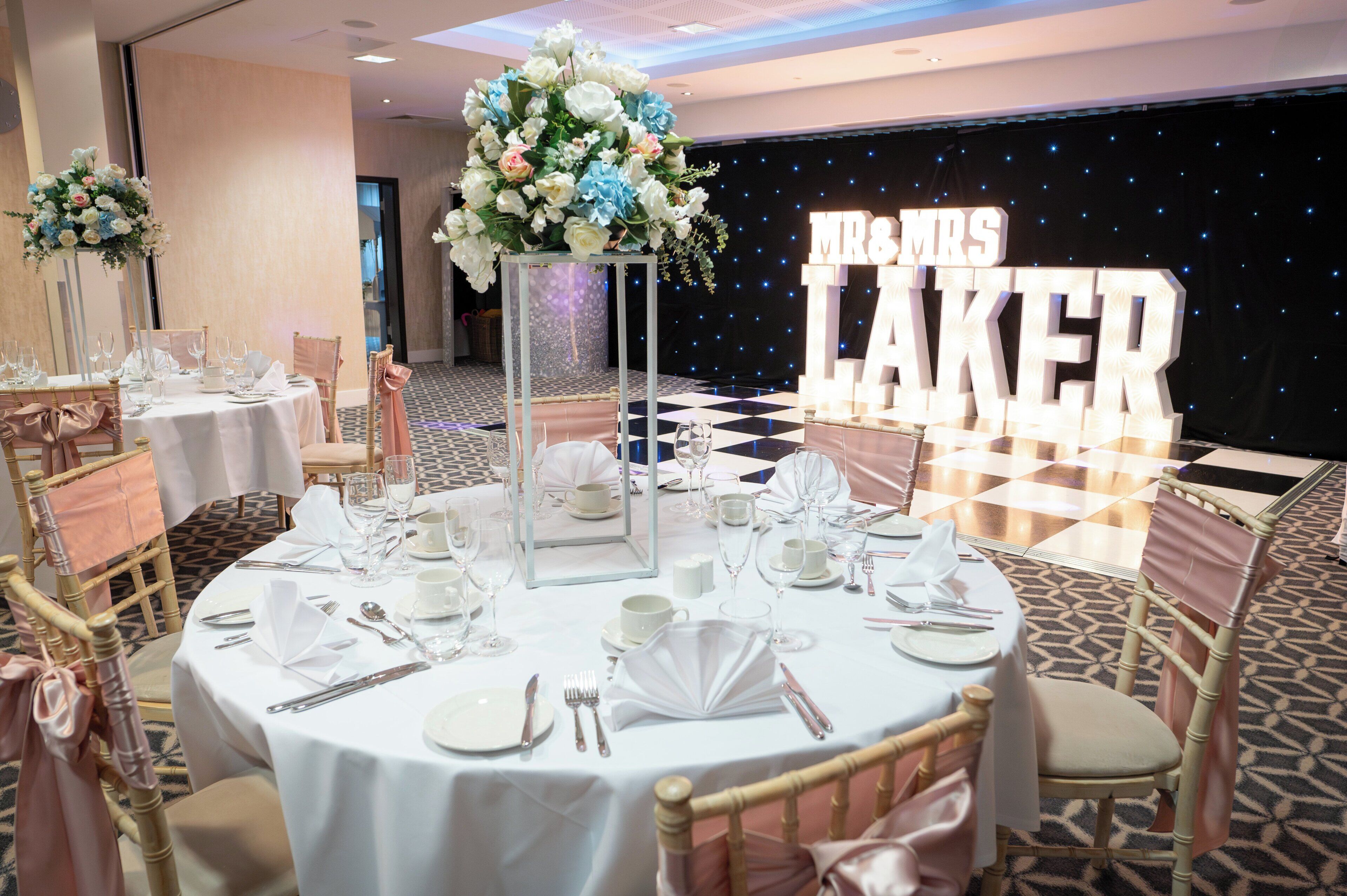 Montaje para una recepción de bodas con mesas redondas, centros de mesa florales y un fondo con letras iluminadas que dicen 'MR & MRS LAKER'.