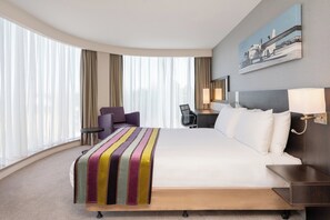 Une chambre d’hôtel avec un grand lit, un bureau, une chaise et une vue sur la ville grâce à de grandes fenêtres.