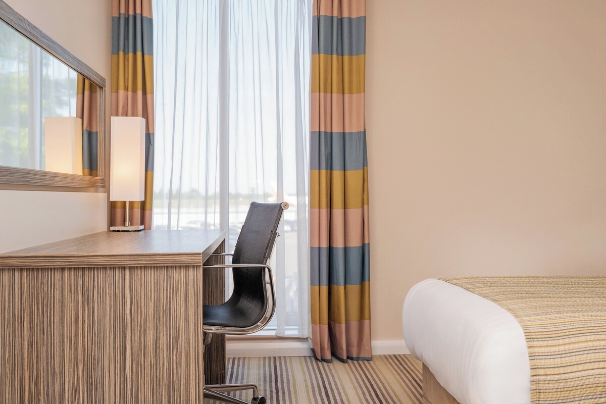 A hotel room with a wooden desk, a chair, a bed, a window with curtains, and a lamp.