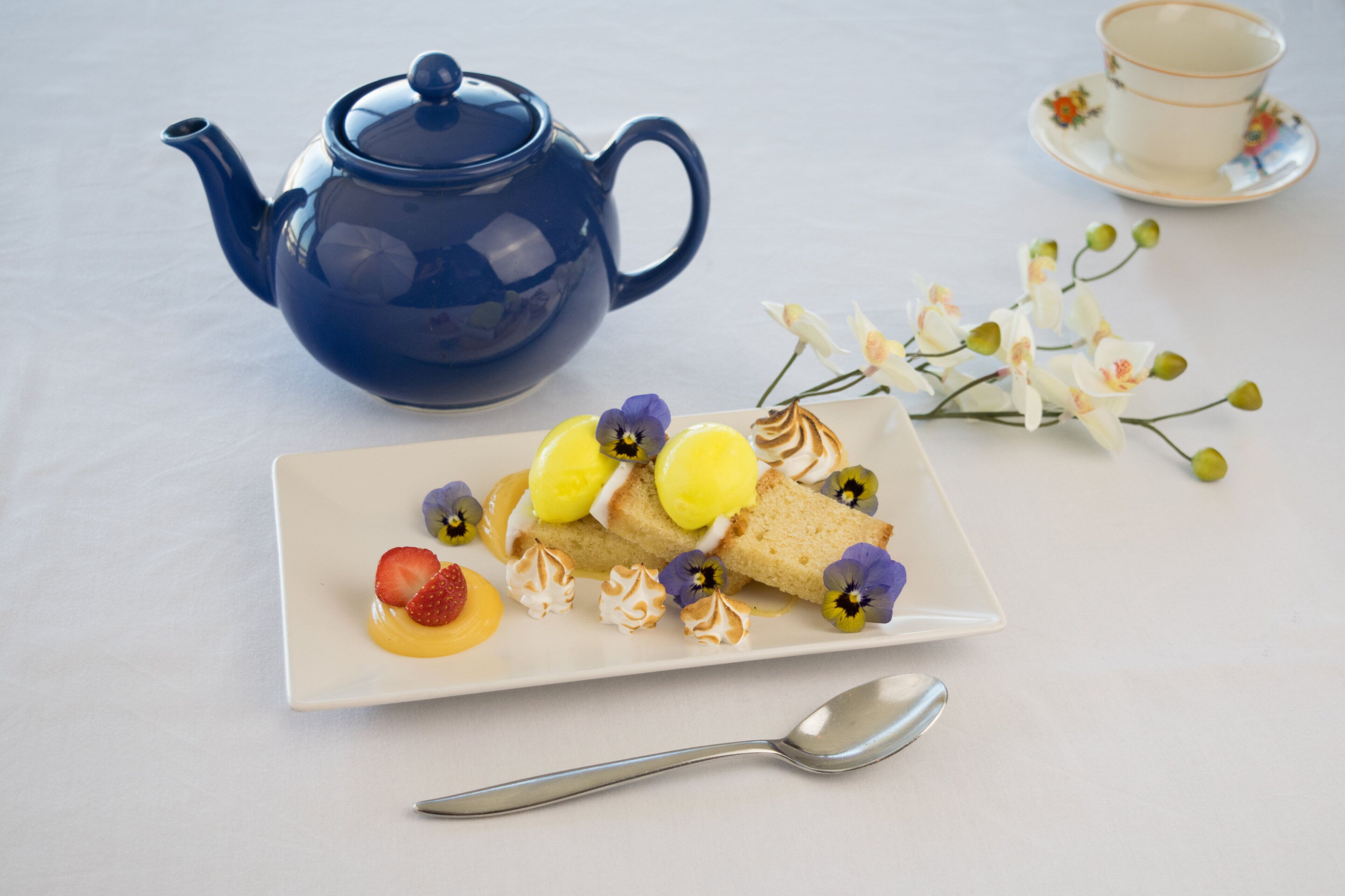 A white plate with a dessert, a blue teapot, a cup, and a spoon on a white table.