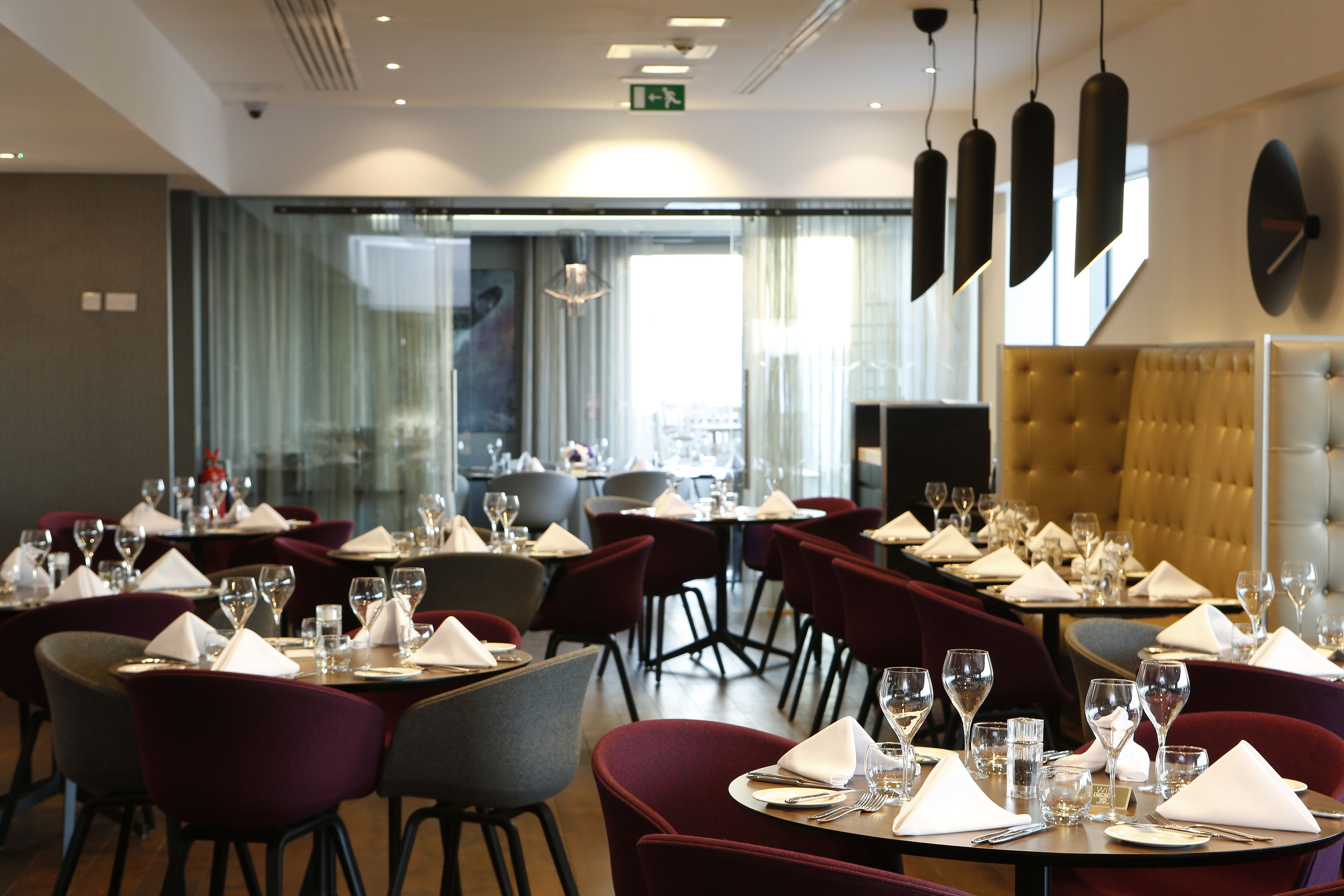 A restaurant with tables set for dining, featuring a mix of red and grey chairs, and a large, tufted gold headboard in the background.