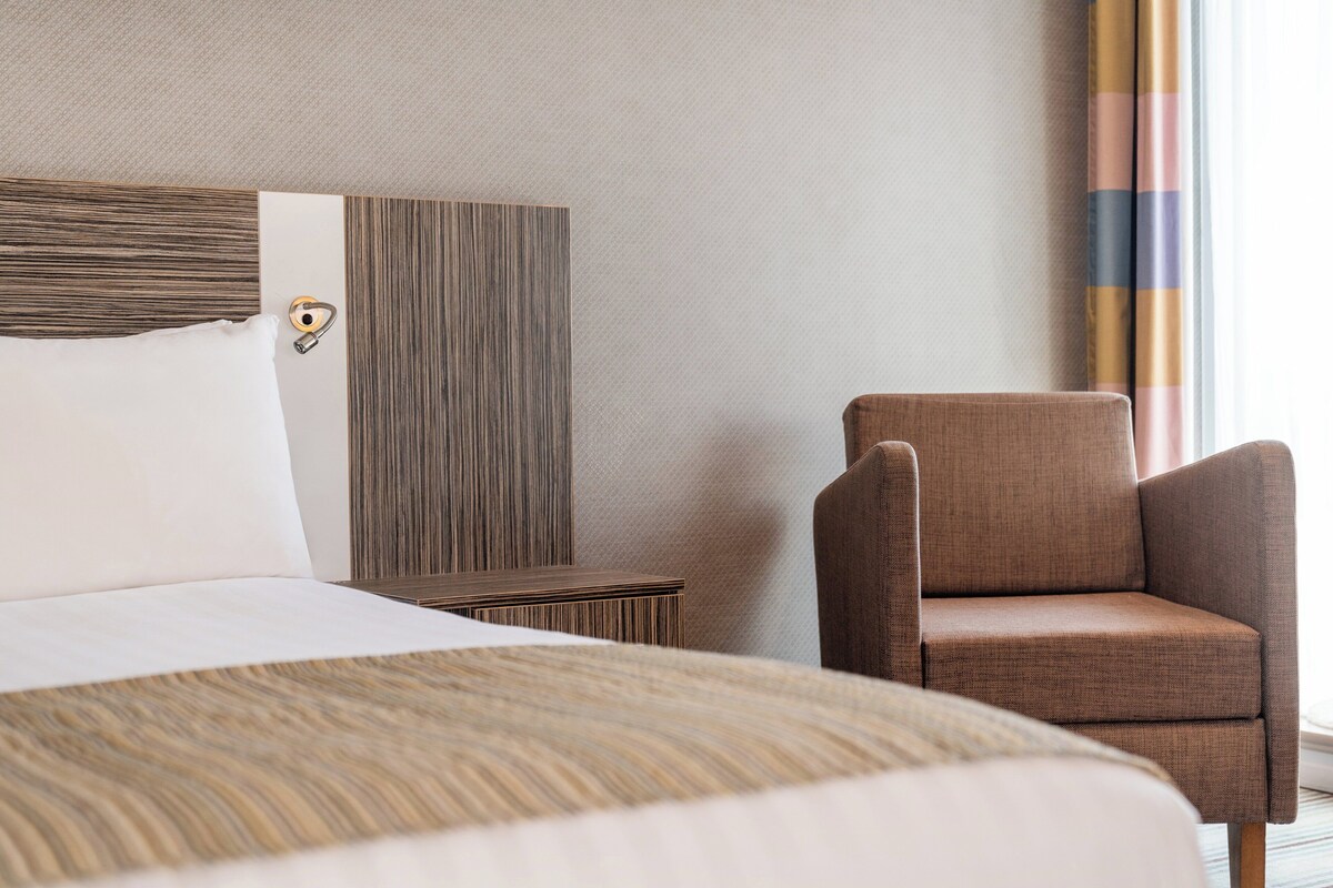 A hotel room with a bed, a brown armchair, a nightstand, and a wall with vertical lines.