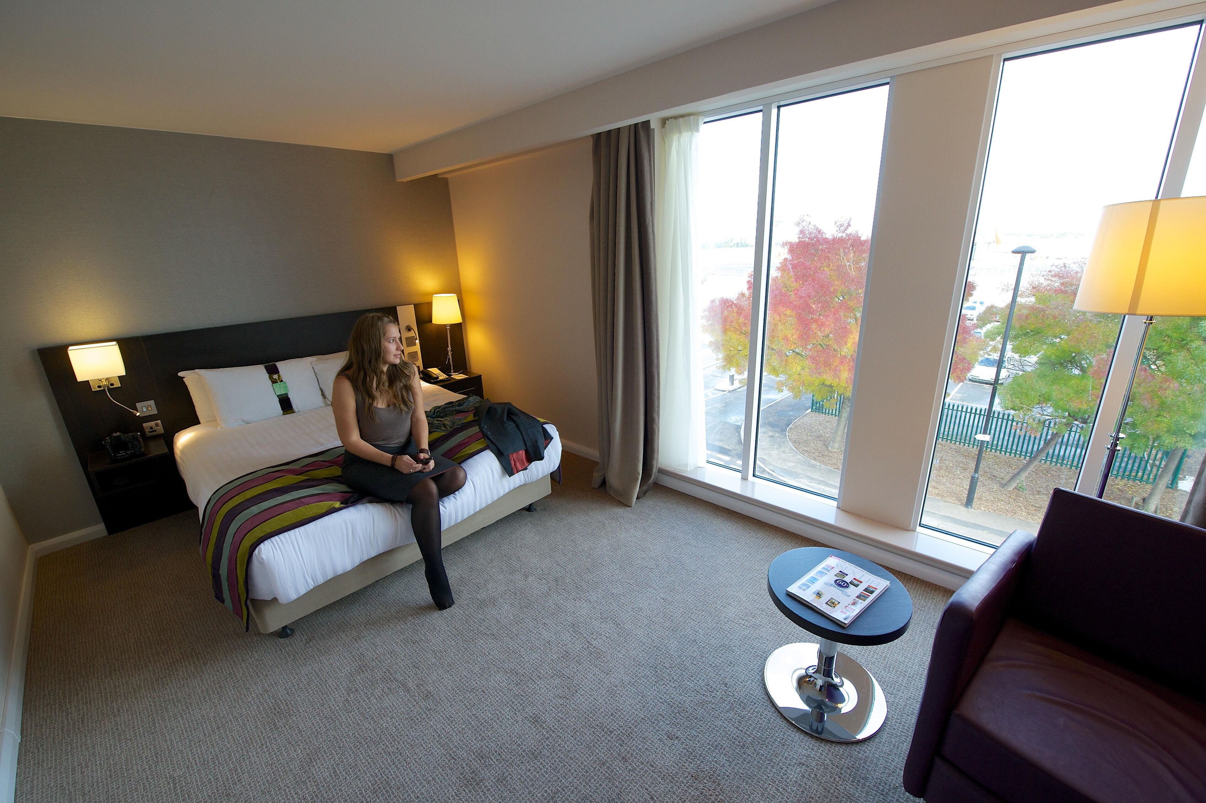 A hotel room with a bed, bedside tables, a small table with a magazine, and large windows with a view of trees.