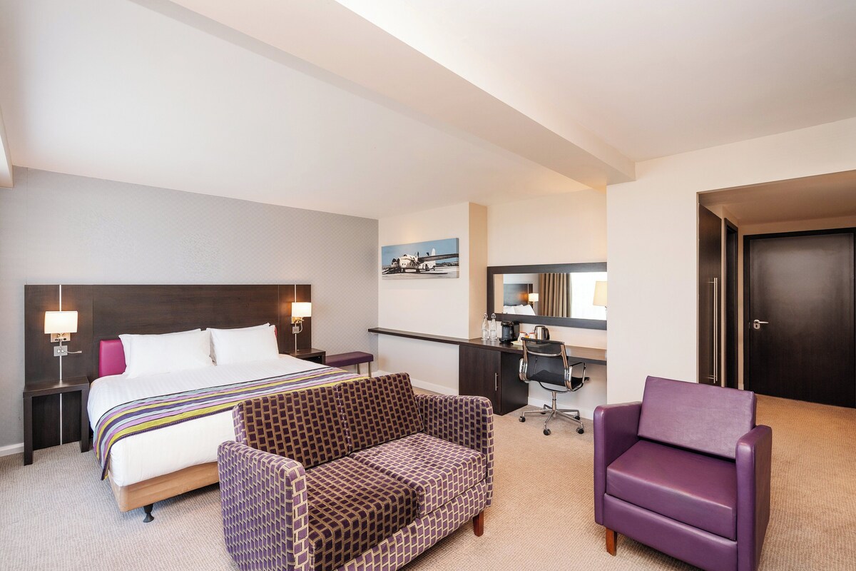 A hotel room with a large bed, a desk, a purple armchair, and a patterned sofa.