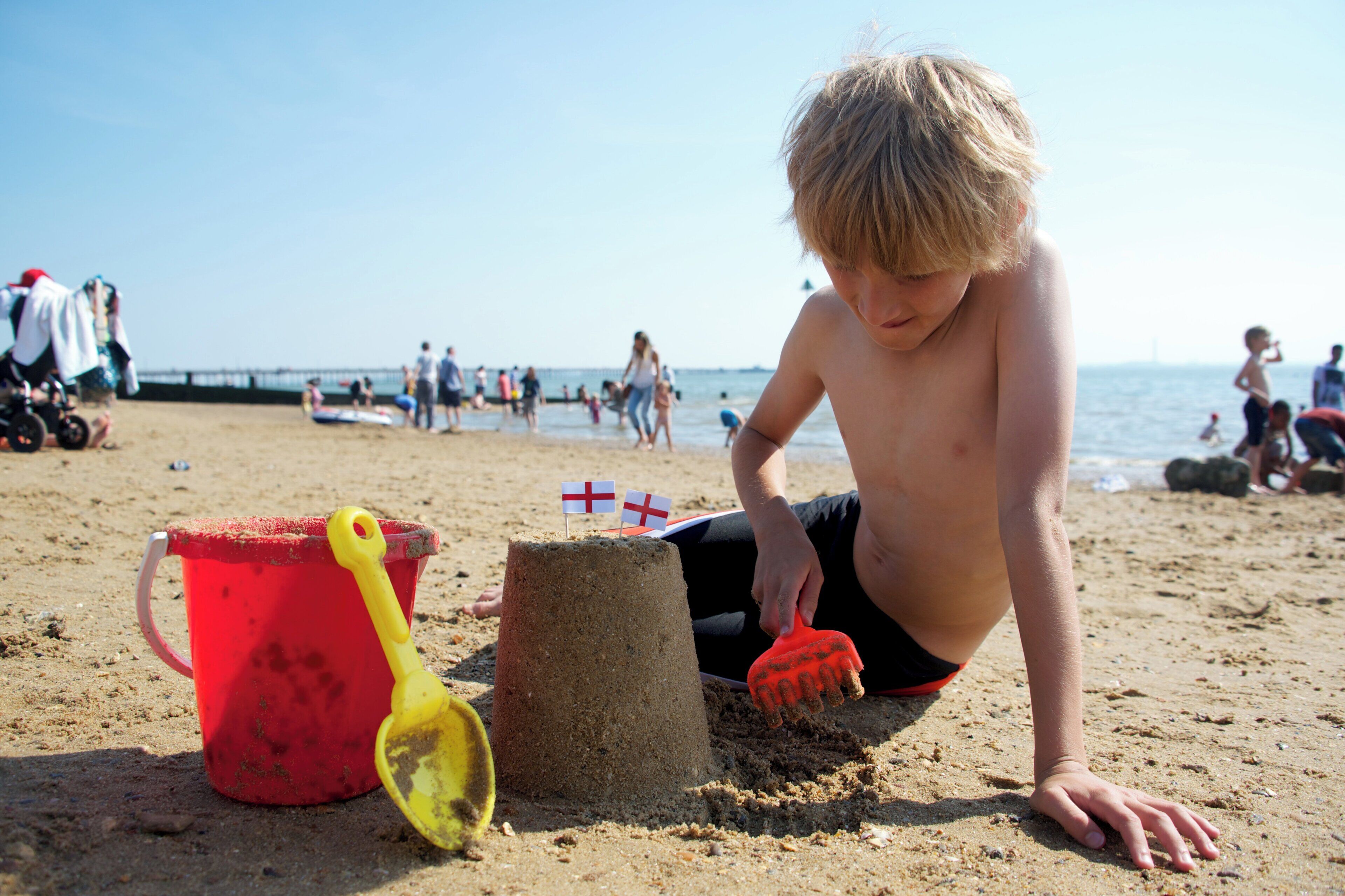 A beach scene with a child building a sandcastle, a red bucket, and a yellow shovel.