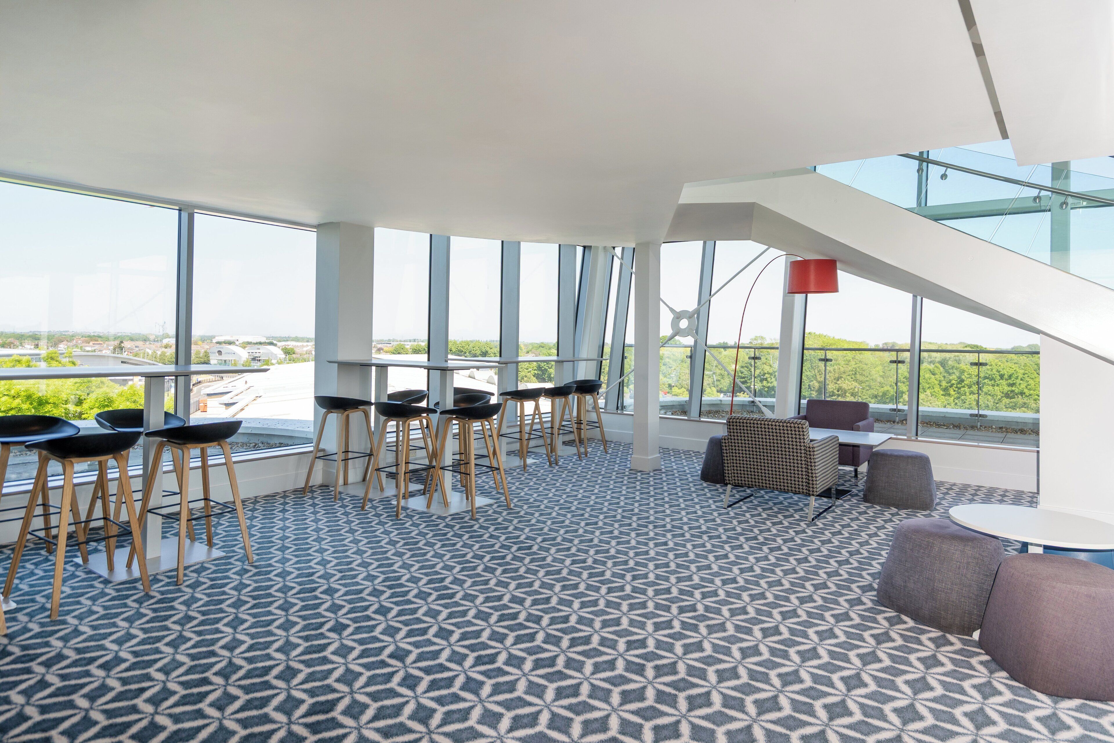 A modern interior with a geometric patterned floor, a staircase, and large windows offering a view of greenery and buildings.