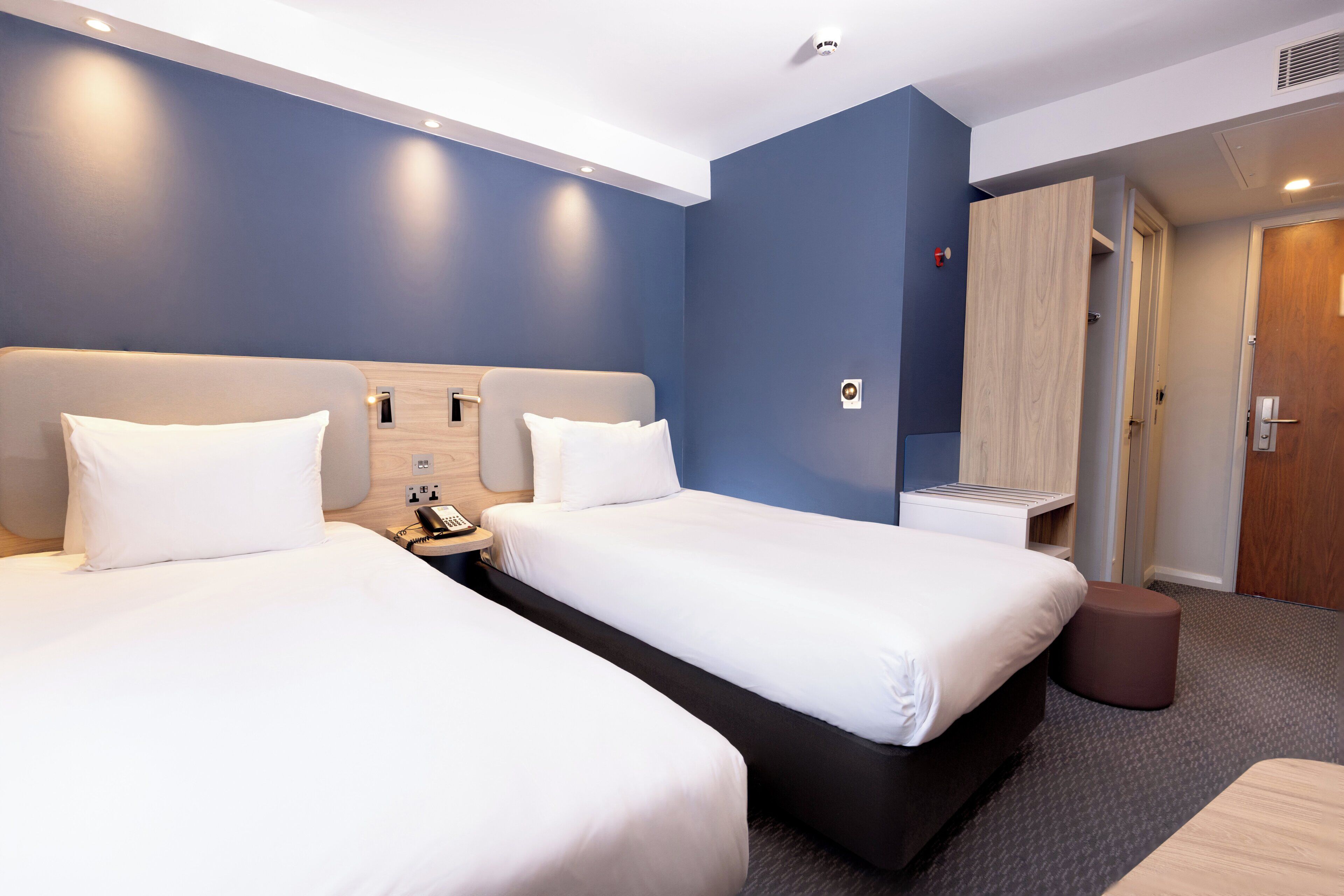 A hotel room with two beds, a wooden wardrobe, a small stool, and a bedside table with a phone.
