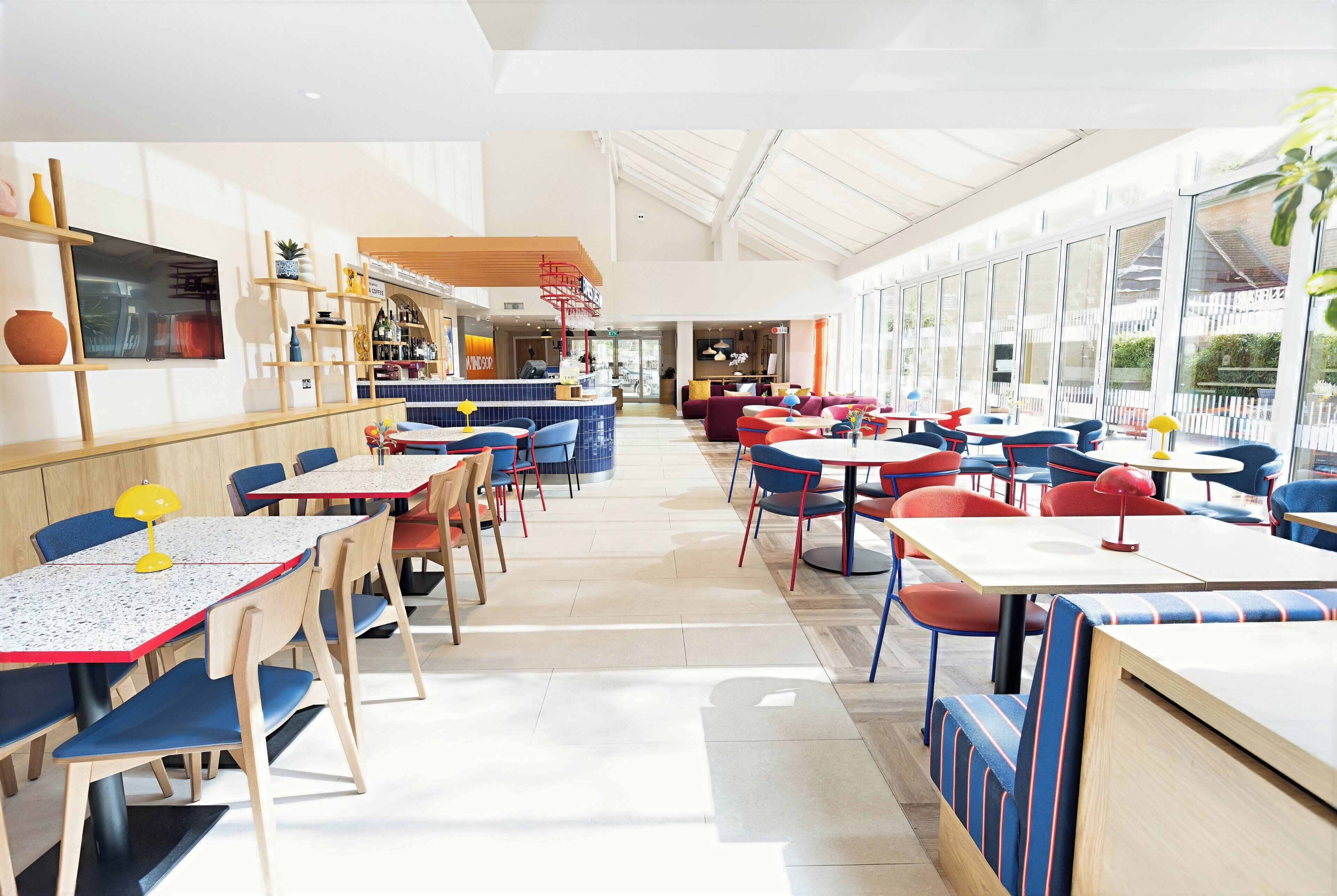 Un comedor amplio e interior con varias mesas y sillas, una zona de bar con estanterías, y grandes ventanales que permiten la entrada de luz natural.