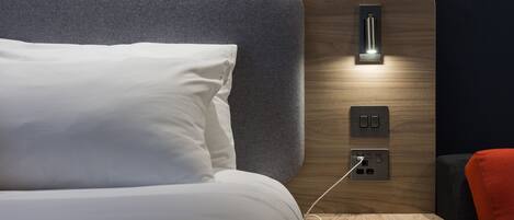 Una cama bien hecha con un almohadón blanco y una mesita de noche con un cable de carga y un teléfono inteligente.