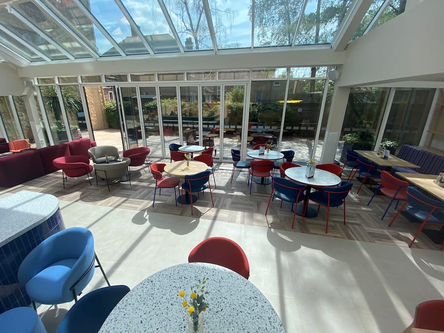 A spacious indoor area with multiple tables and chairs, large windows, and a glass ceiling allowing natural light.