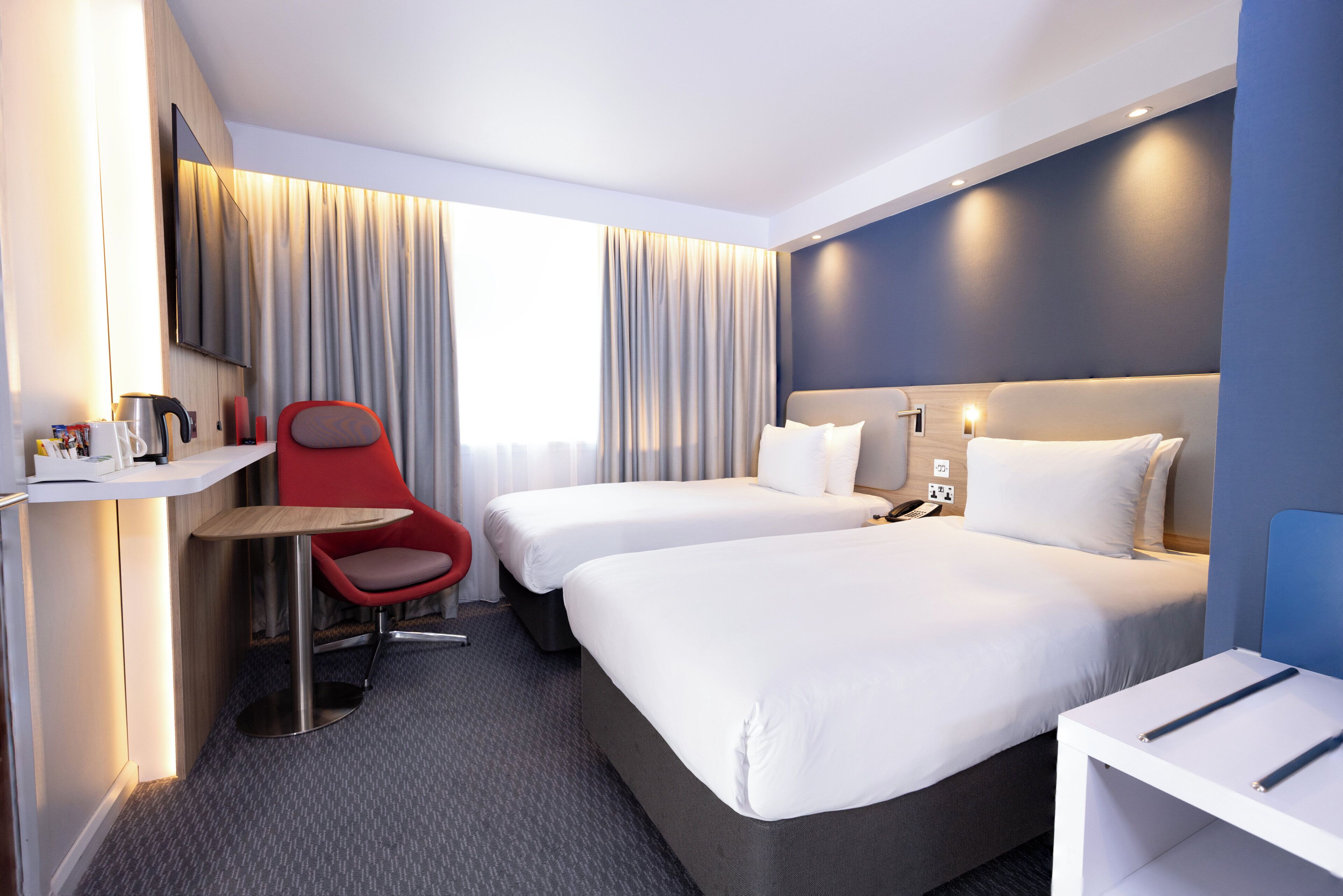 A hotel room with two beds, a desk with a chair, a TV, and a window with curtains.