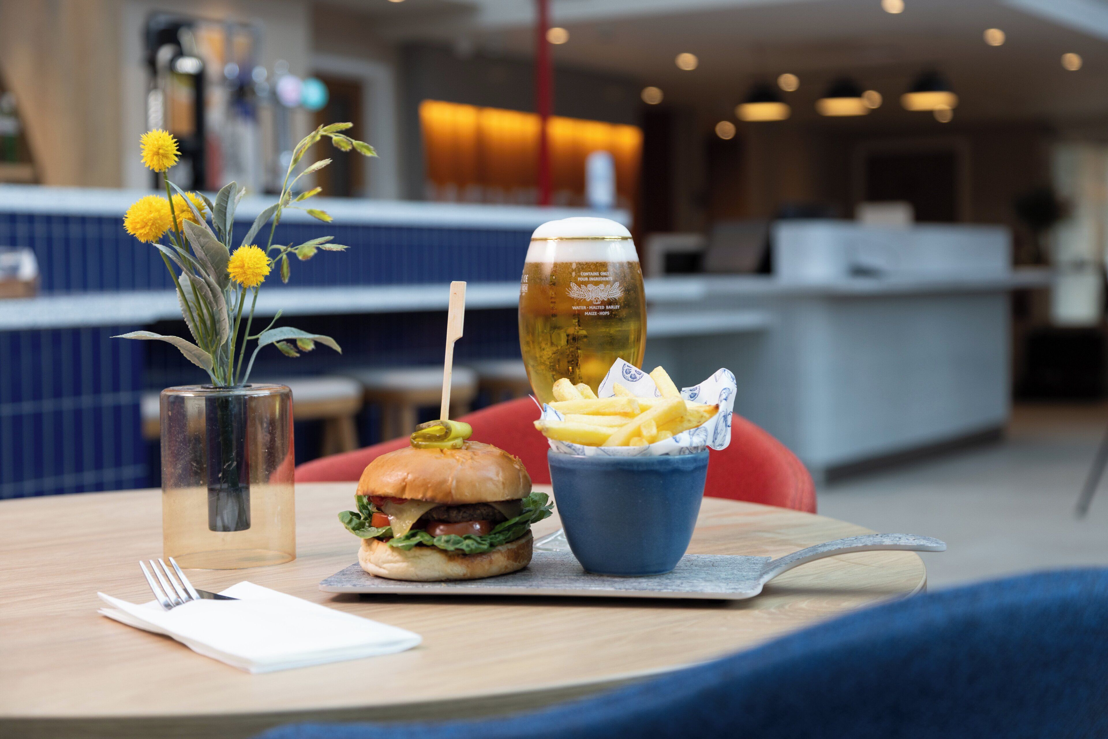 Una mesa de comedor con una hamburguesa, papas fritas, una jarra de cerveza y un florero con flores.