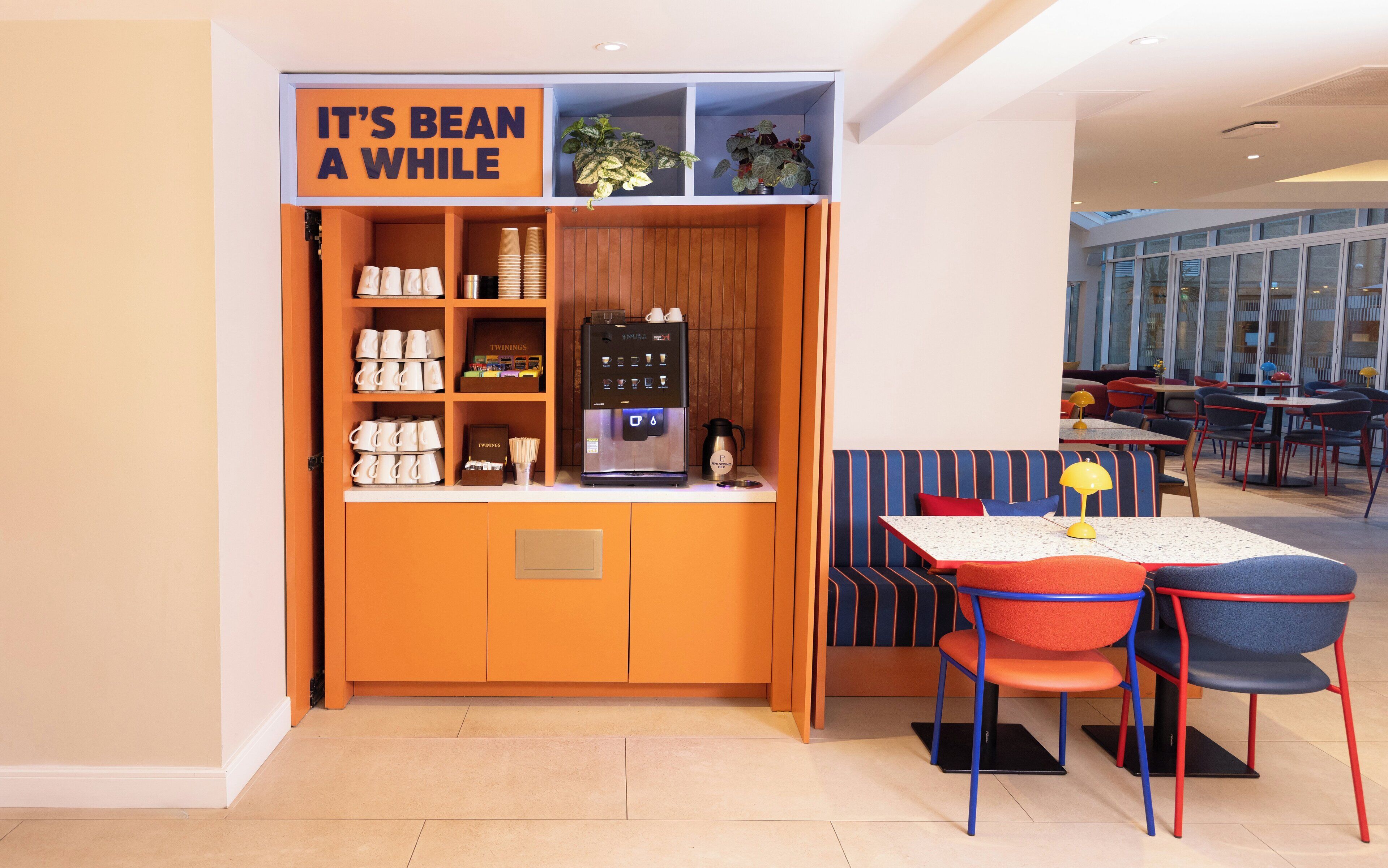 A coffee station with a sign reading "IT'S BEAN A WHILE", a coffee machine, and seating area with tables and chairs.