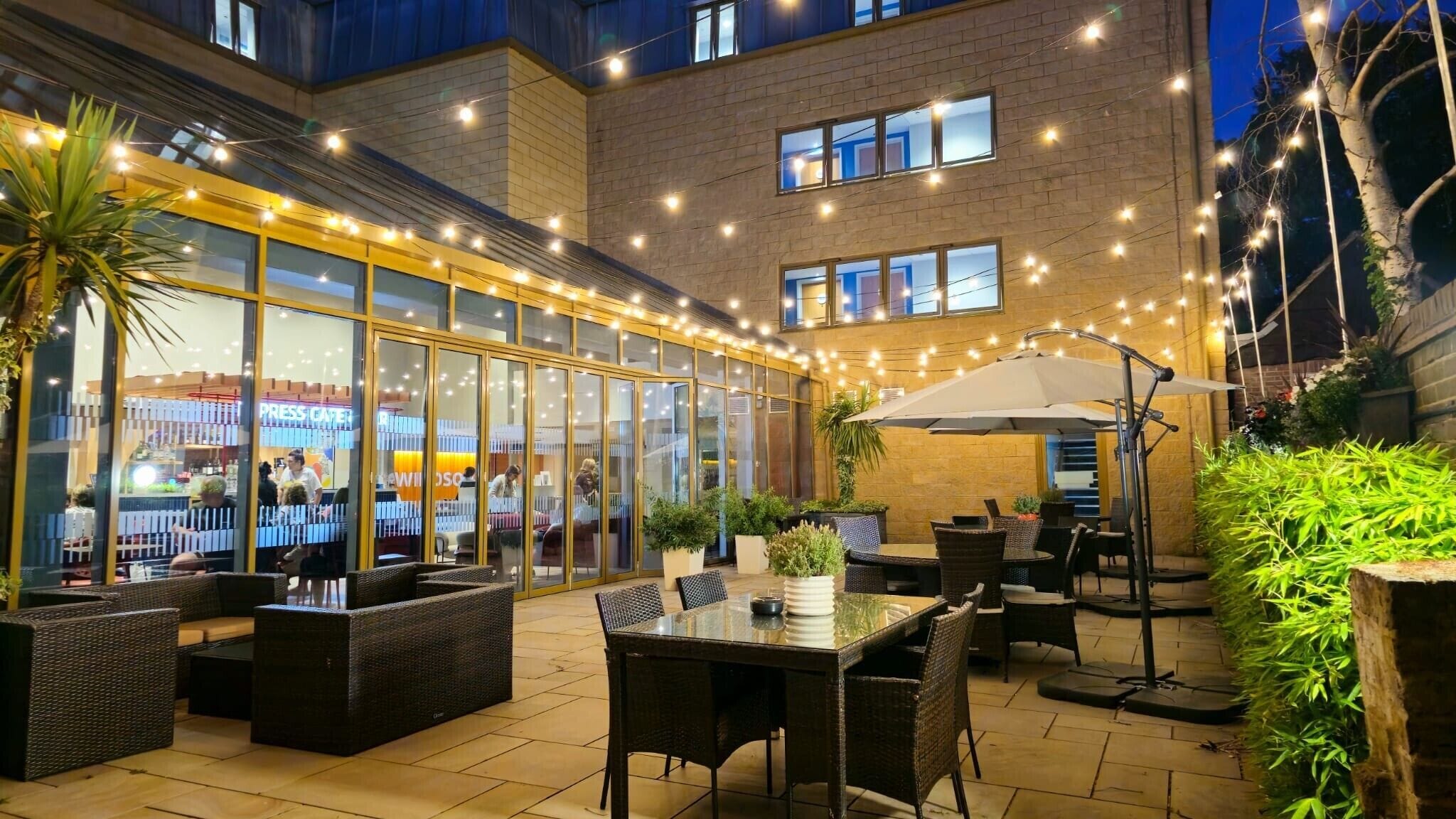 Un espacio al aire libre con patio, mobiliario de mimbre, cerramiento de vidrio y luces colgantes.