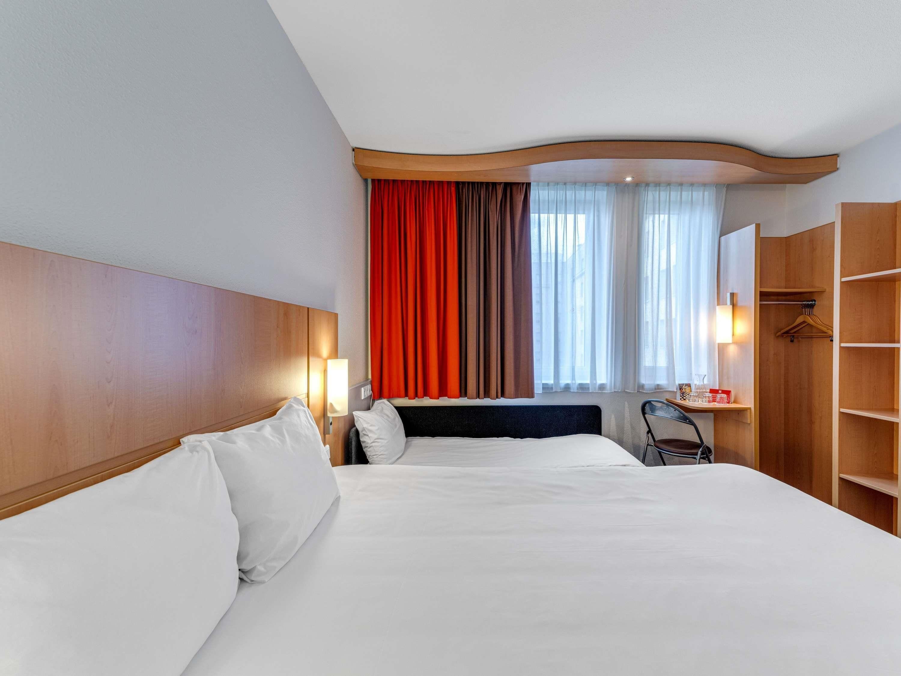 Une chambre d’hôtel avec deux lits, une tête de lit en bois, un bureau avec une chaise, une armoire et une fenêtre avec des rideaux.