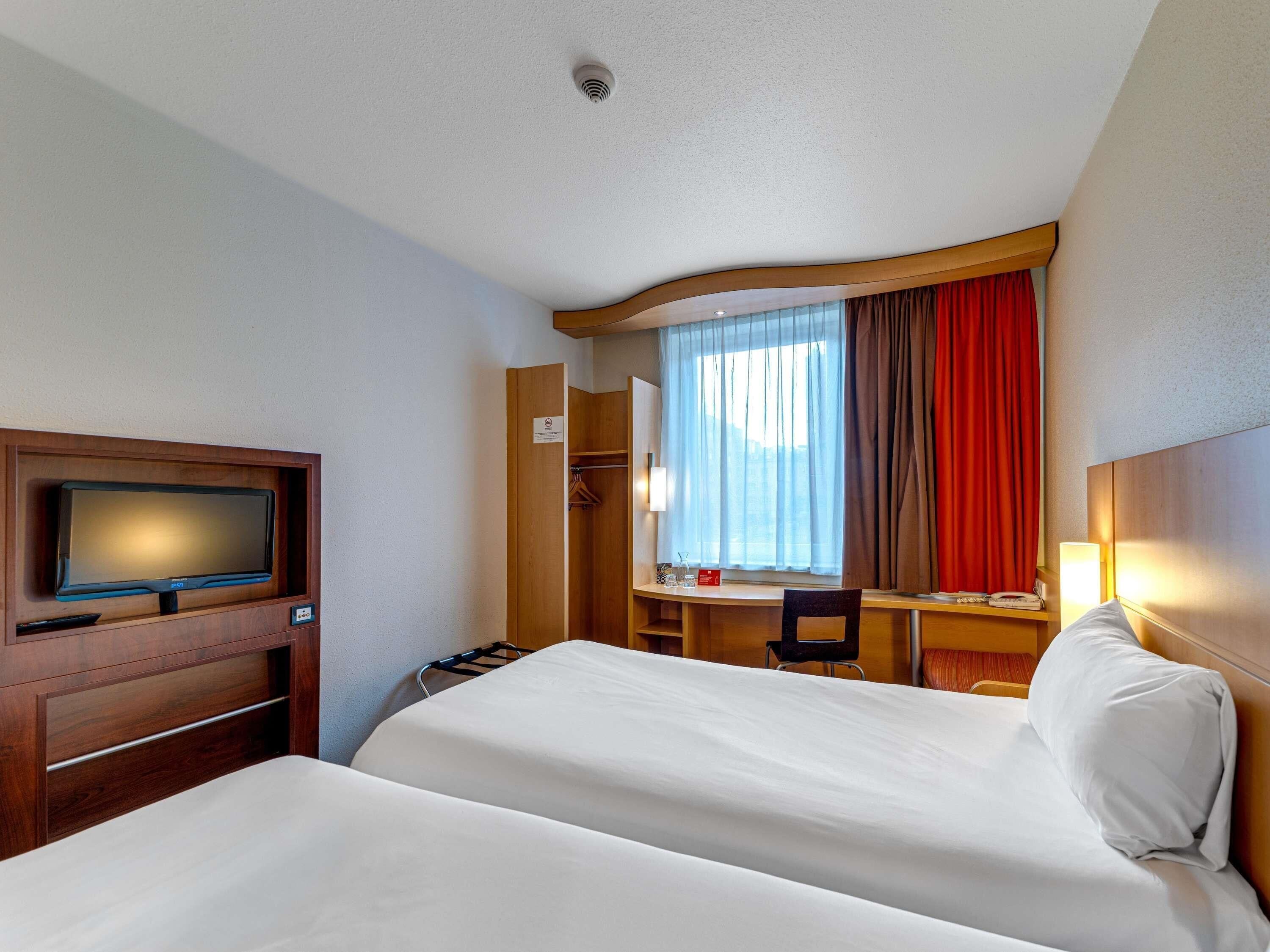 A hotel room with two beds, a desk, a TV, and a window with curtains.