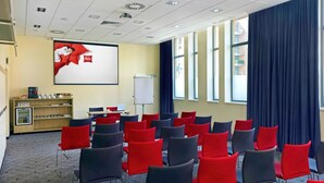A conference room with rows of chairs, a projector screen, a whiteboard, and a coffee station. - ibis Krakow Stare Miasto (Kraków)