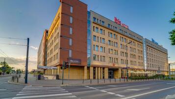 A multi-story hotel with a prominent 'ibis Hotel' sign, a visible entrance, and a pedestrian crossing.