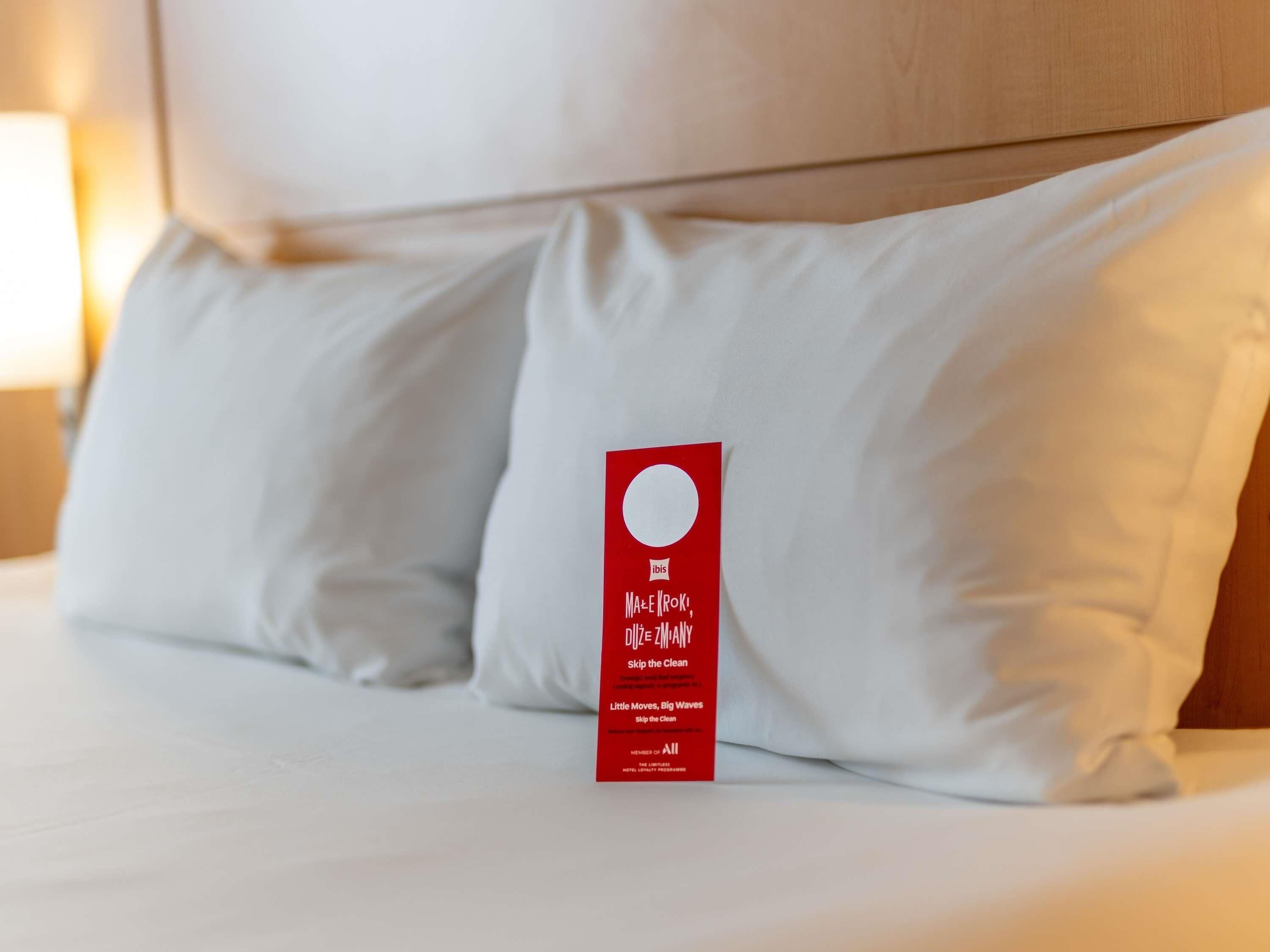 A hotel room with a neatly made bed, white pillows, and a red tag on the bed.