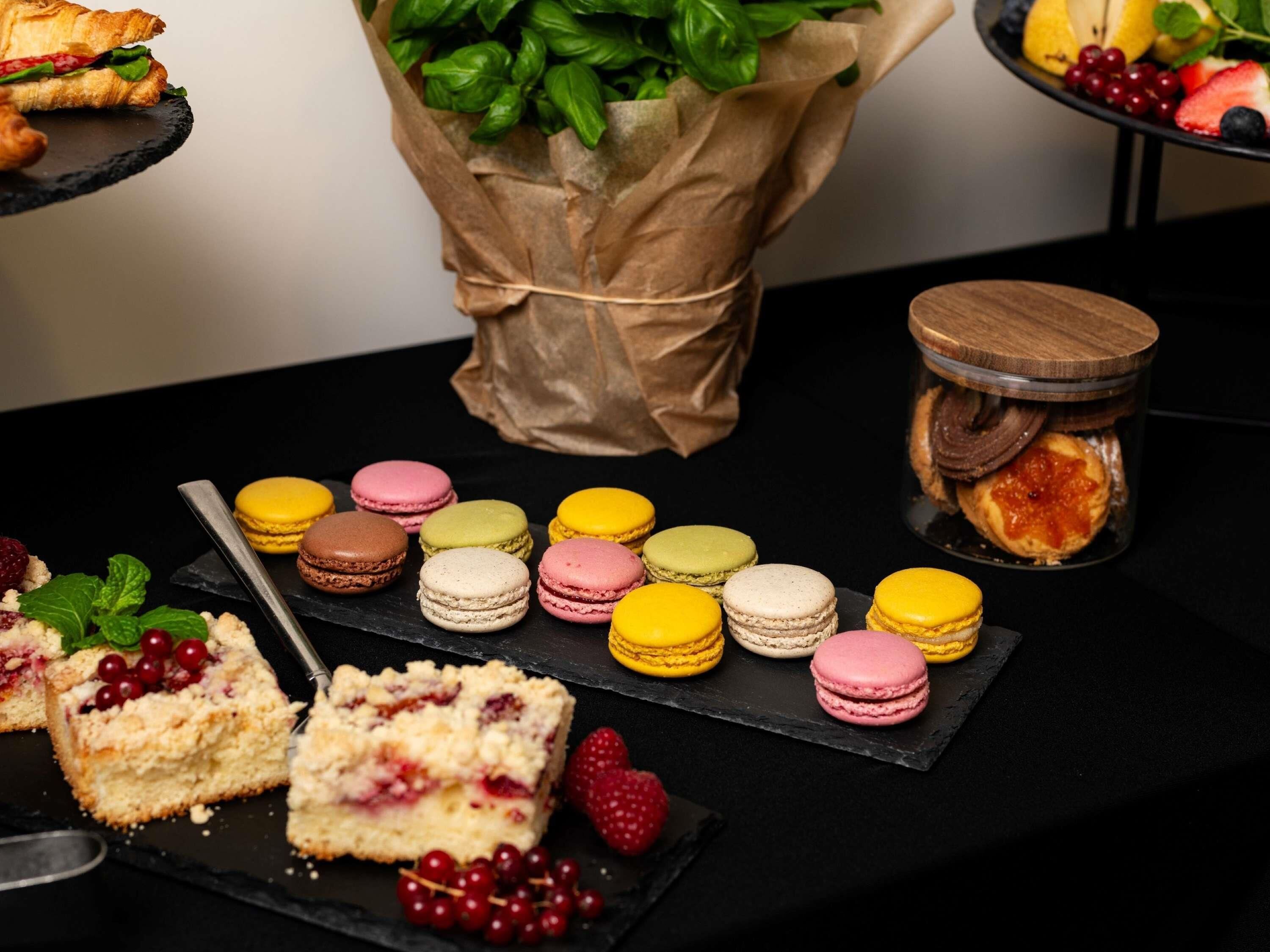 A variety of desserts including macarons, a pastry sandwich, and a fruit platter on a black slate serving board.