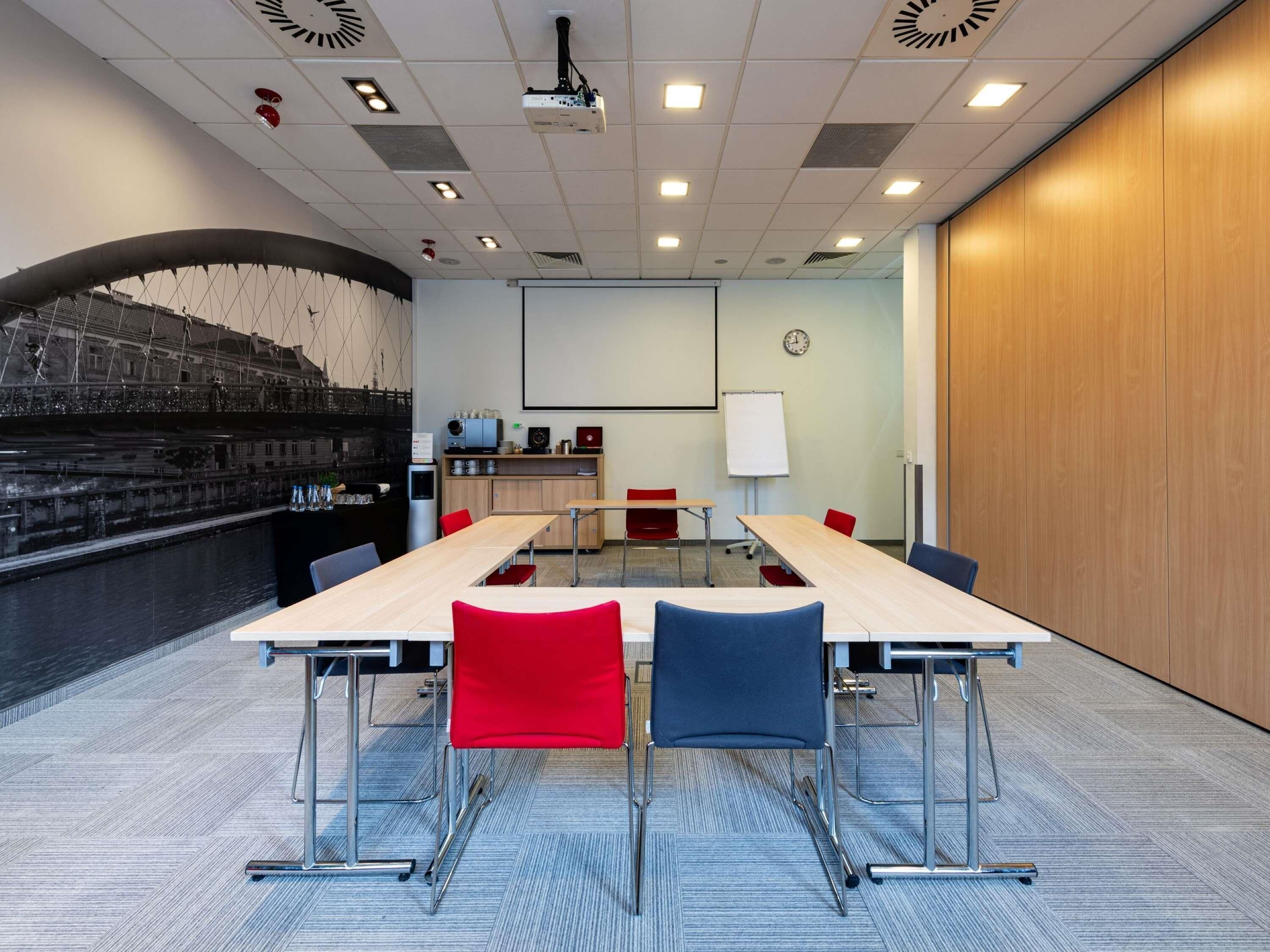 A conference room with a long table, red and blue chairs, a projector, a whiteboard, and a clock on the wall.