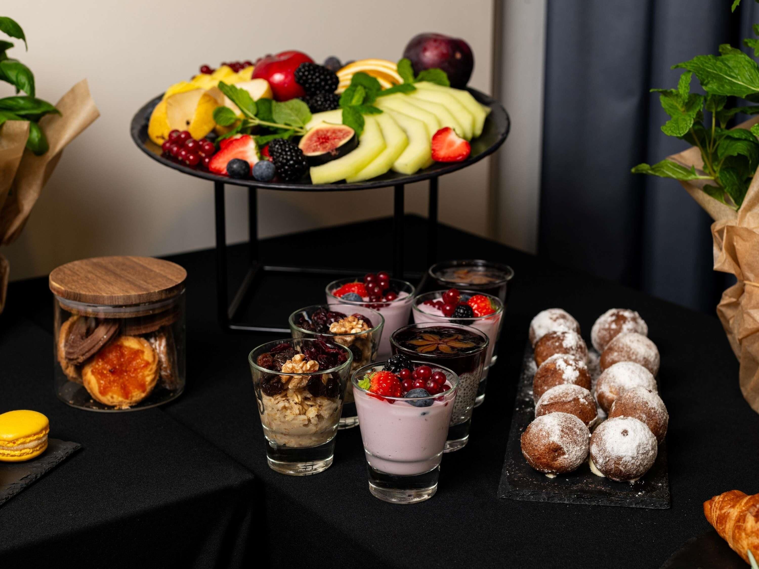 A table with a fruit platter, dessert glasses, a jar of dried fruits, and powdered sugar-covered pastries.