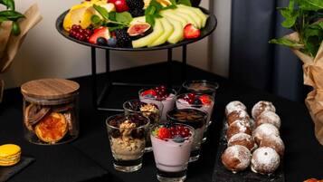 Una mesa con una bandeja de frutas, vasos de postre, un tarro de frutas secas y pasteles cubiertos de azúcar glas.