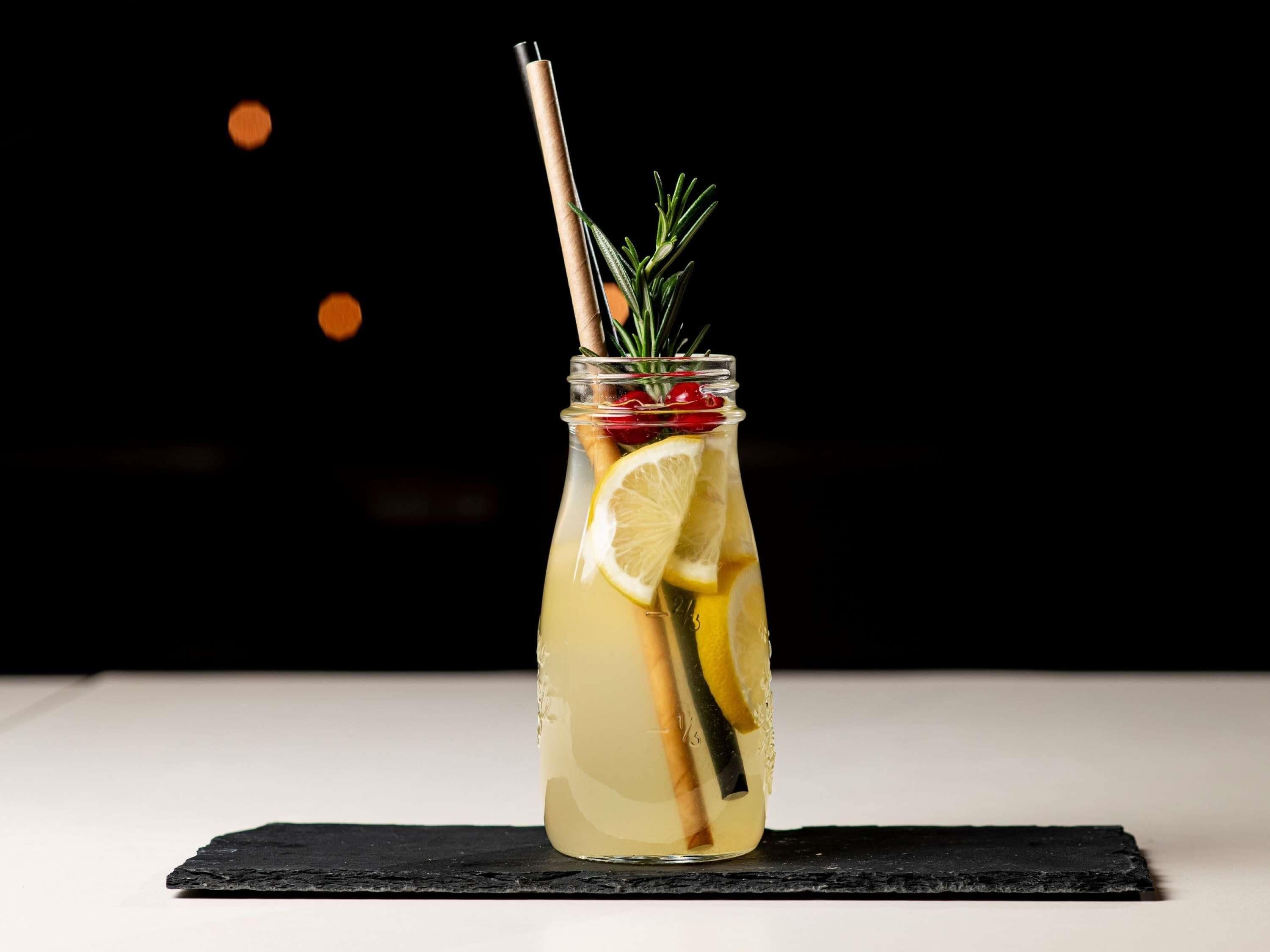 A clear glass bottle with a straw, containing a yellowish liquid, garnished with lemon slices and herbs, placed on a black slate.