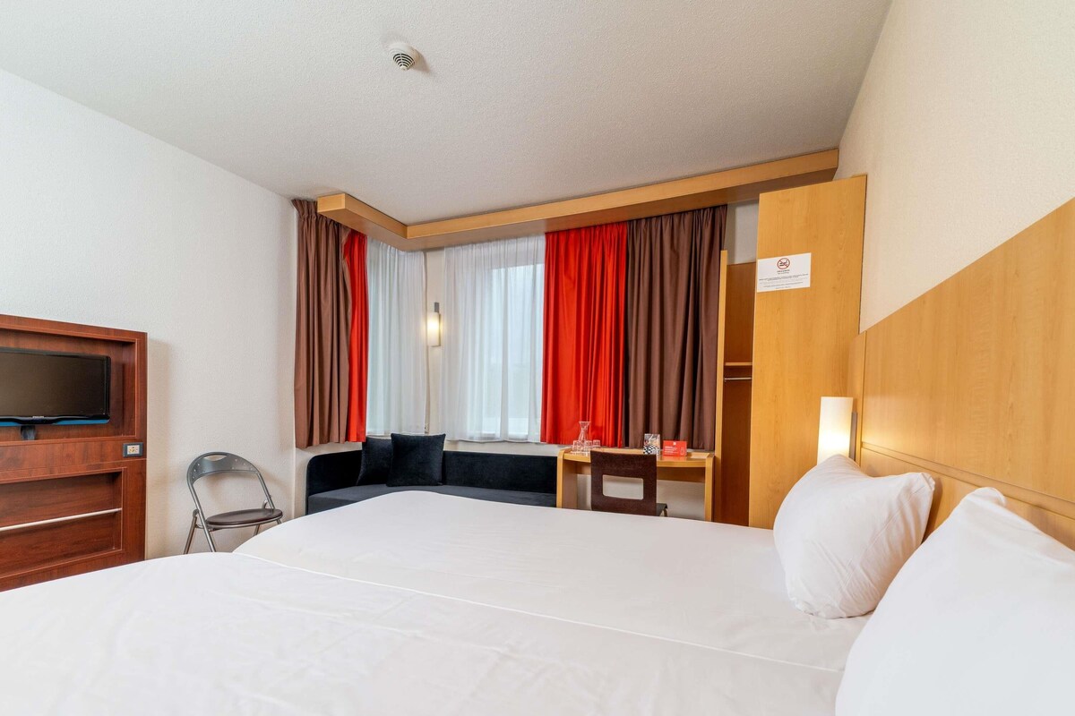 Une chambre d’hôtel avec un grand lit, un bureau avec une chaise, une télévision et une fenêtre avec des rideaux.