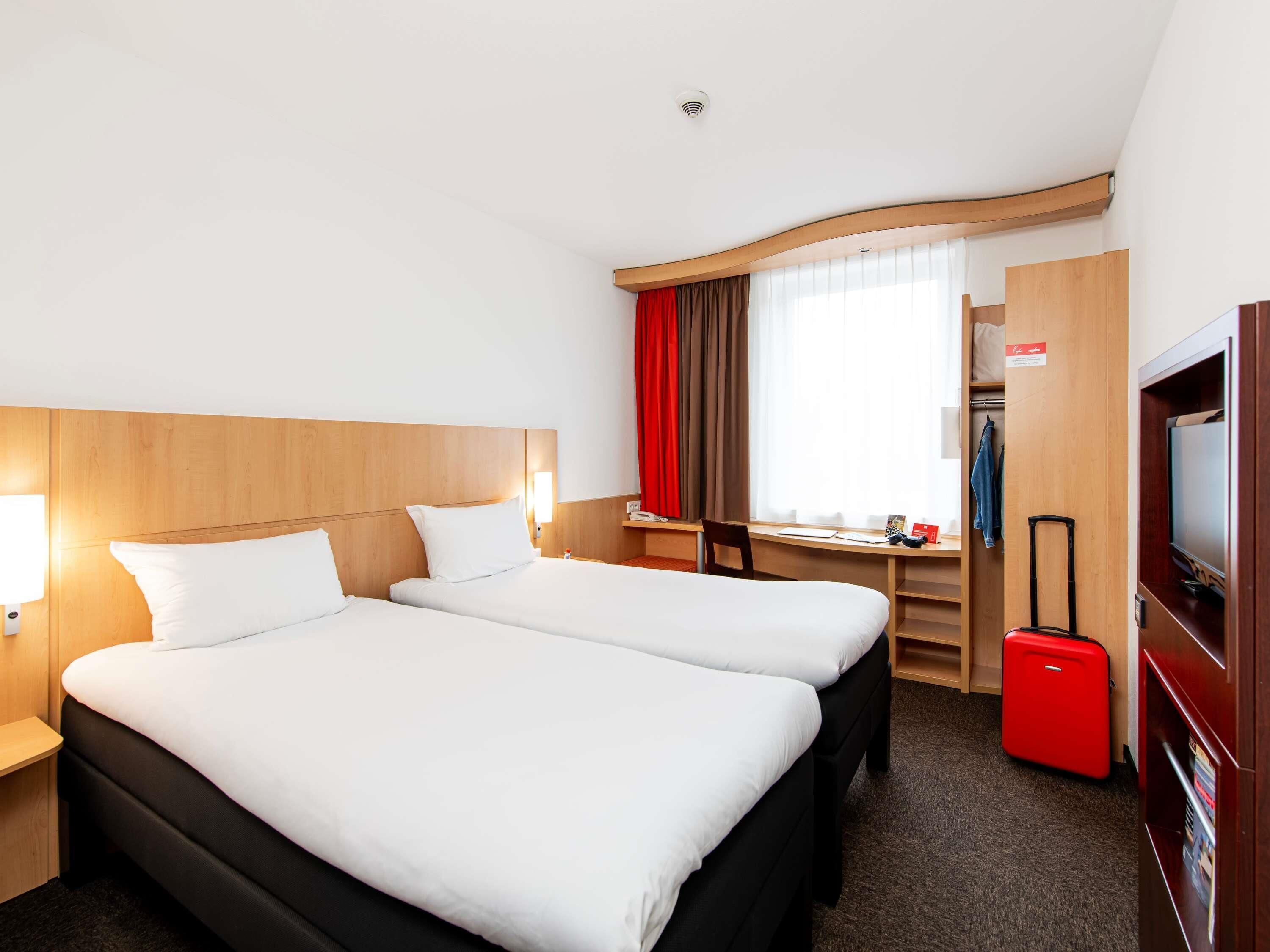 Habitación de hotel con dos camas, un escritorio, un televisor y una maleta roja.
