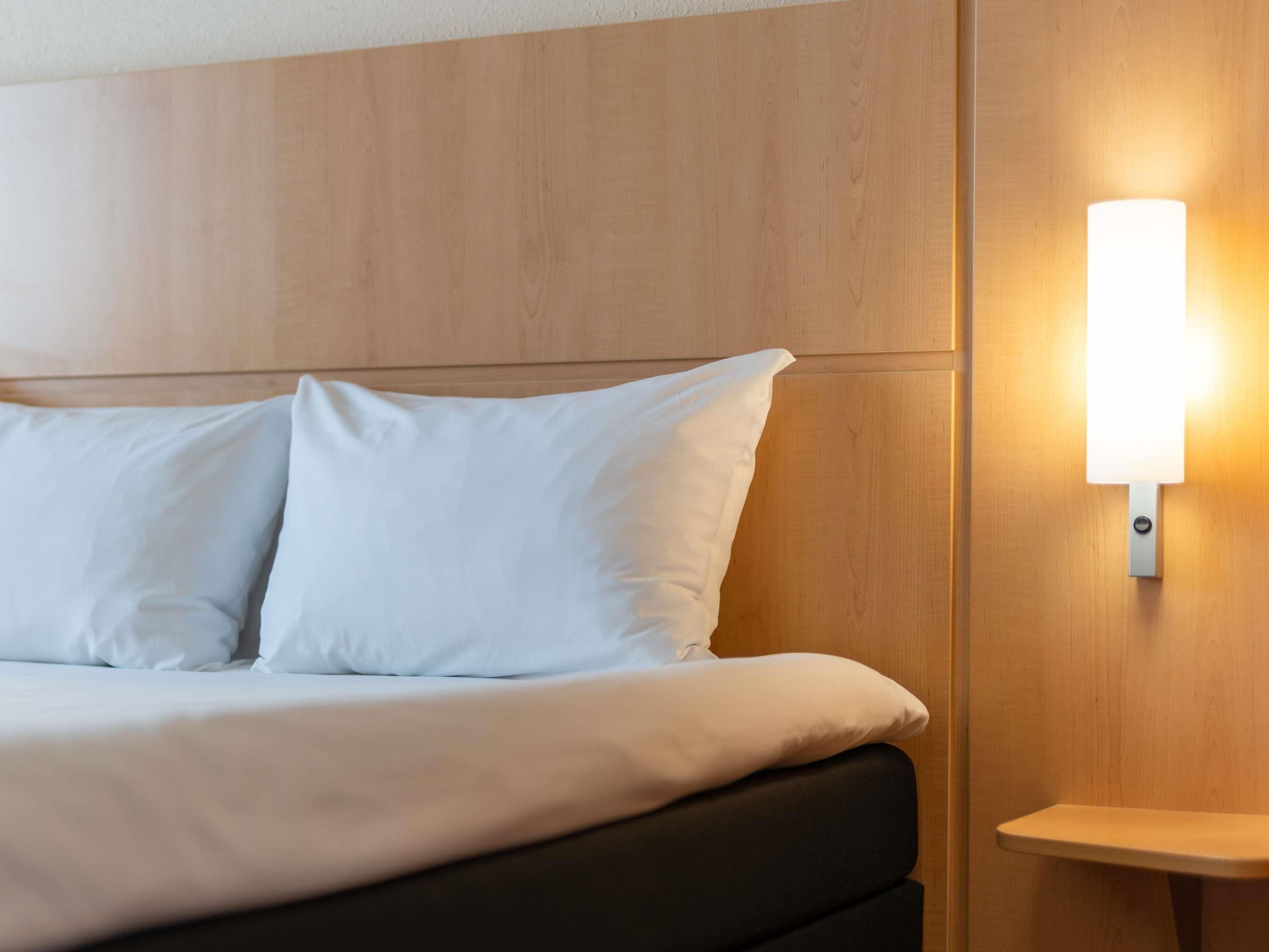 Een bed met witte lakens en een nachtkastje met een wandlamp.