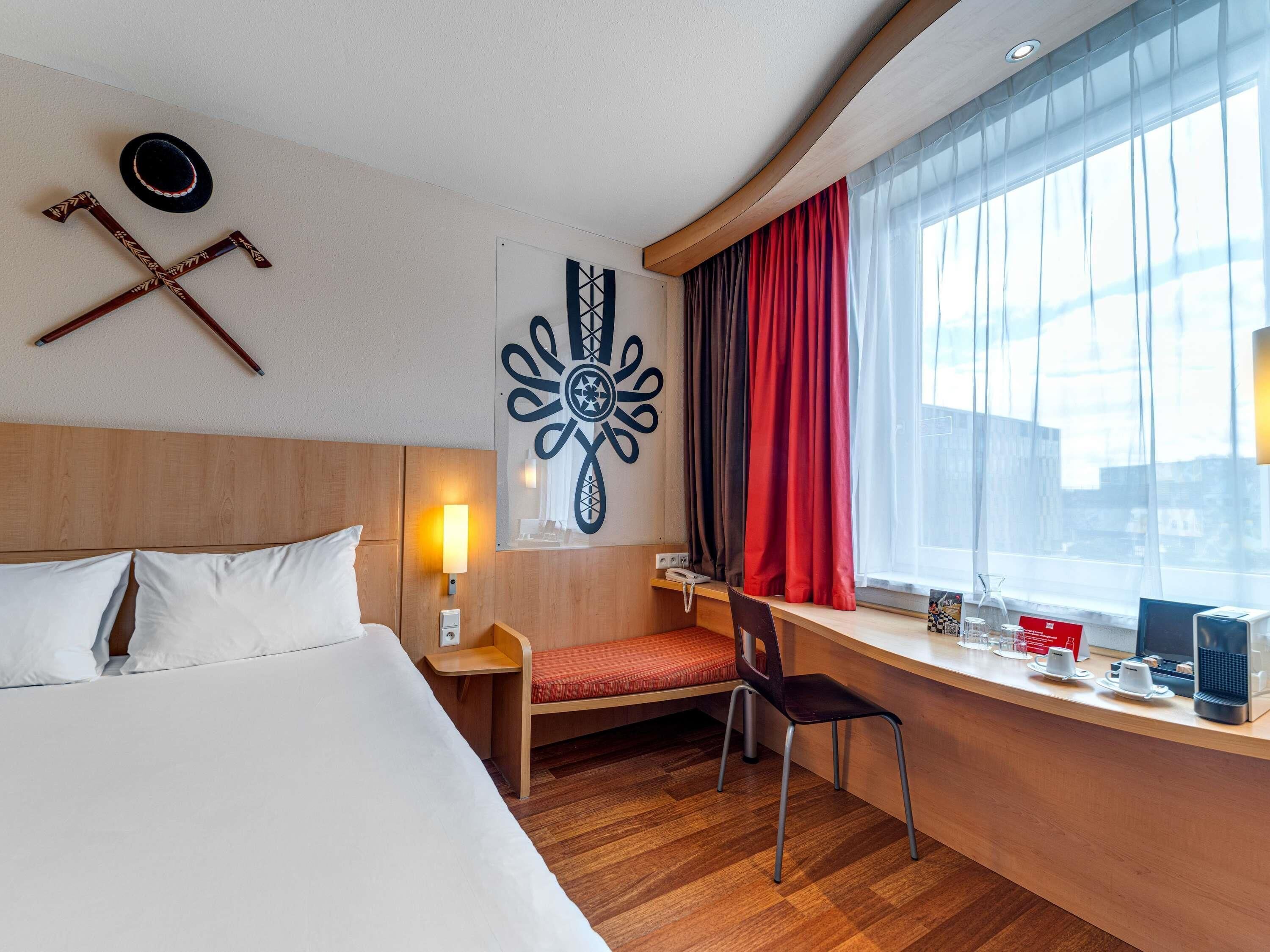 Habitación de hotel con cama, escritorio con silla, lámpara, ventana con cortinas y decoración en la pared.