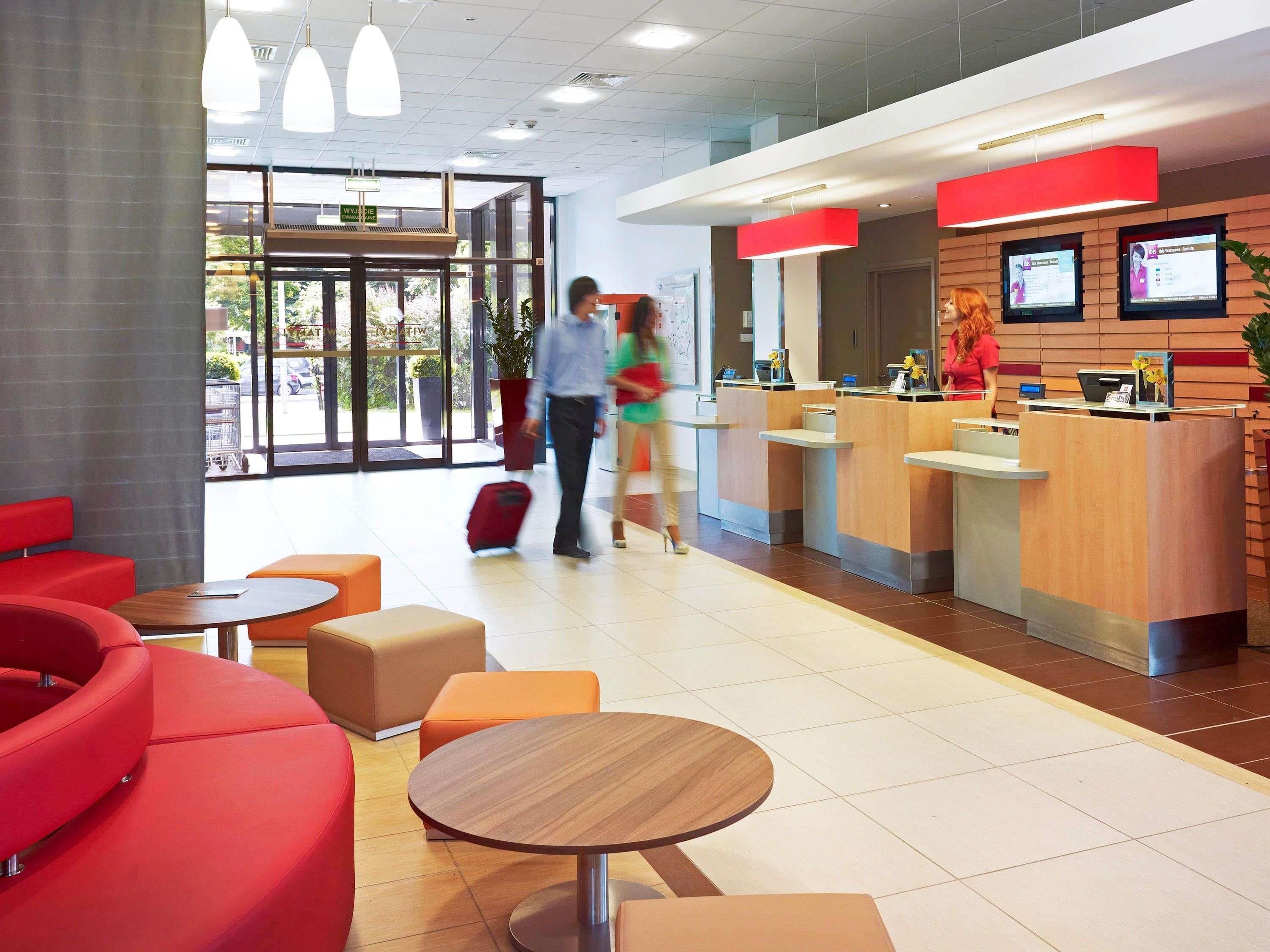 A modern reception area with a check-in desk, seating area with red and orange chairs, and a large window providing natural light.