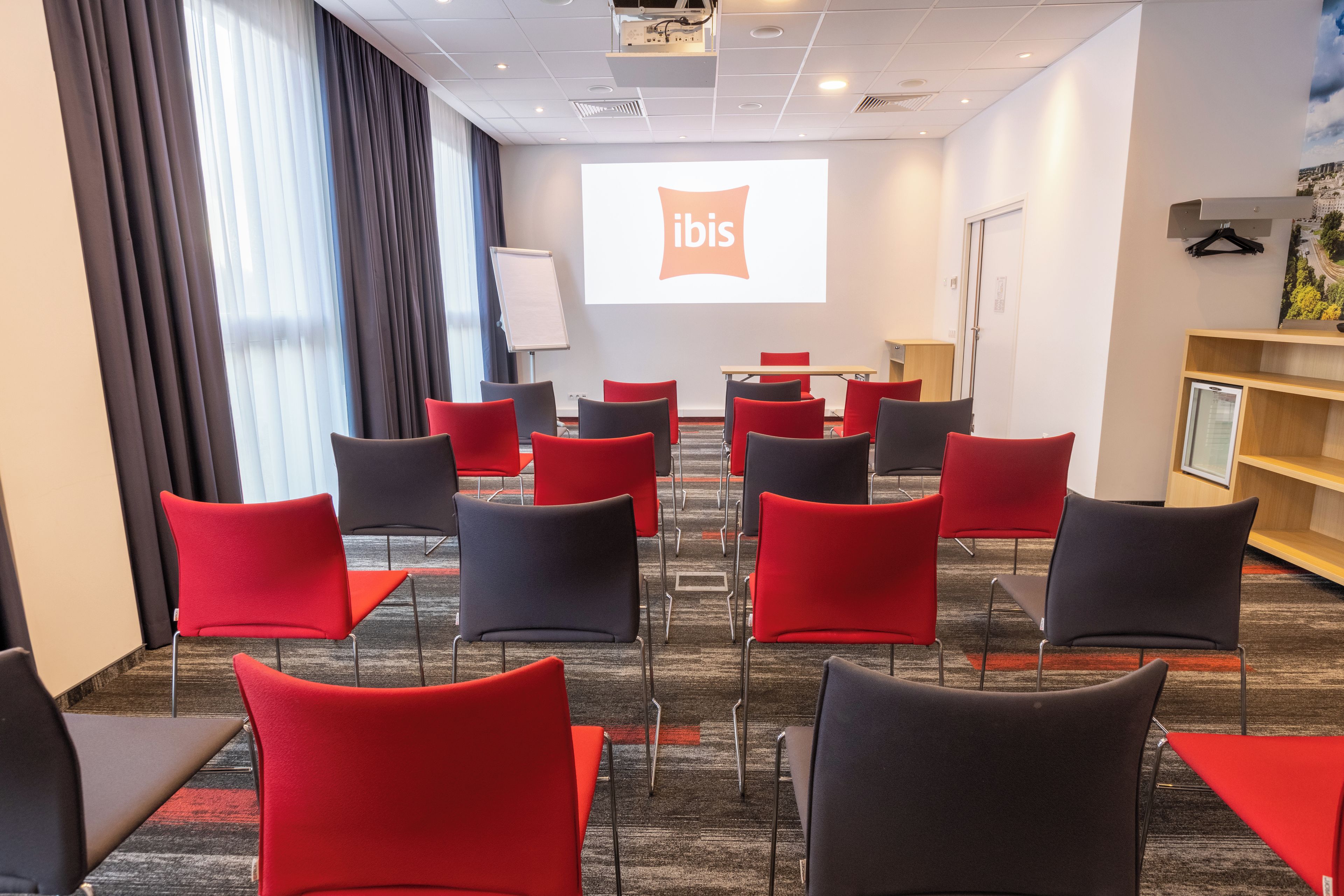 A conference room with rows of red and black chairs, a projector screen displaying 'ibis', and a whiteboard.
